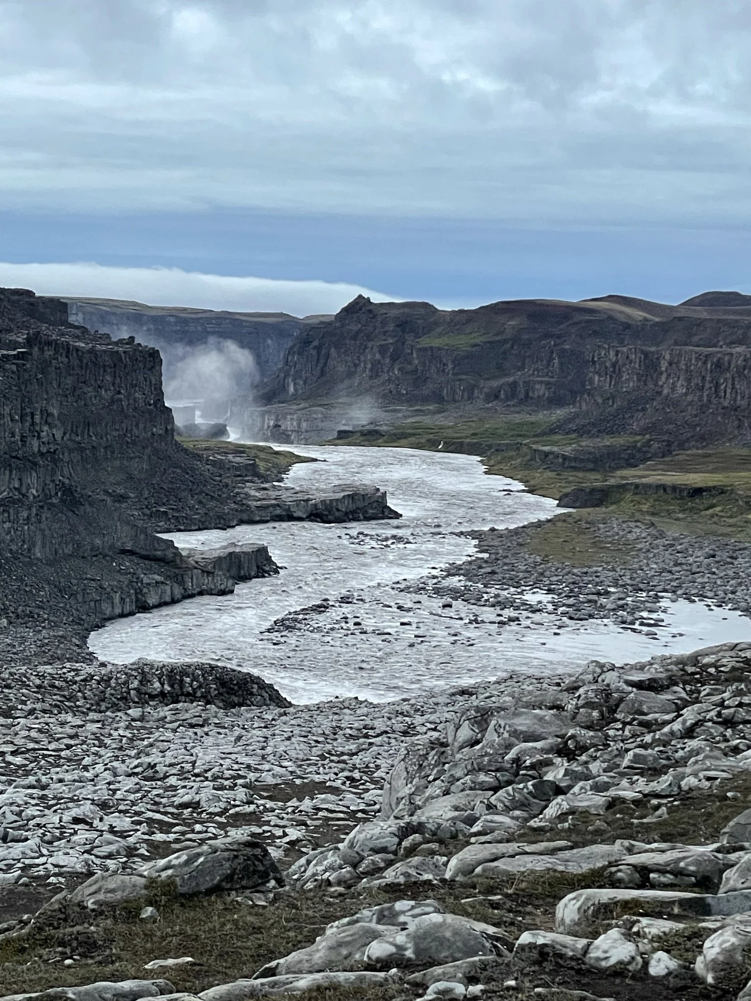 A rugged canyon landscape with a river flowing through rocky terrain, cliffs on either side, and a mist rising in the distance under a cloudy sky.