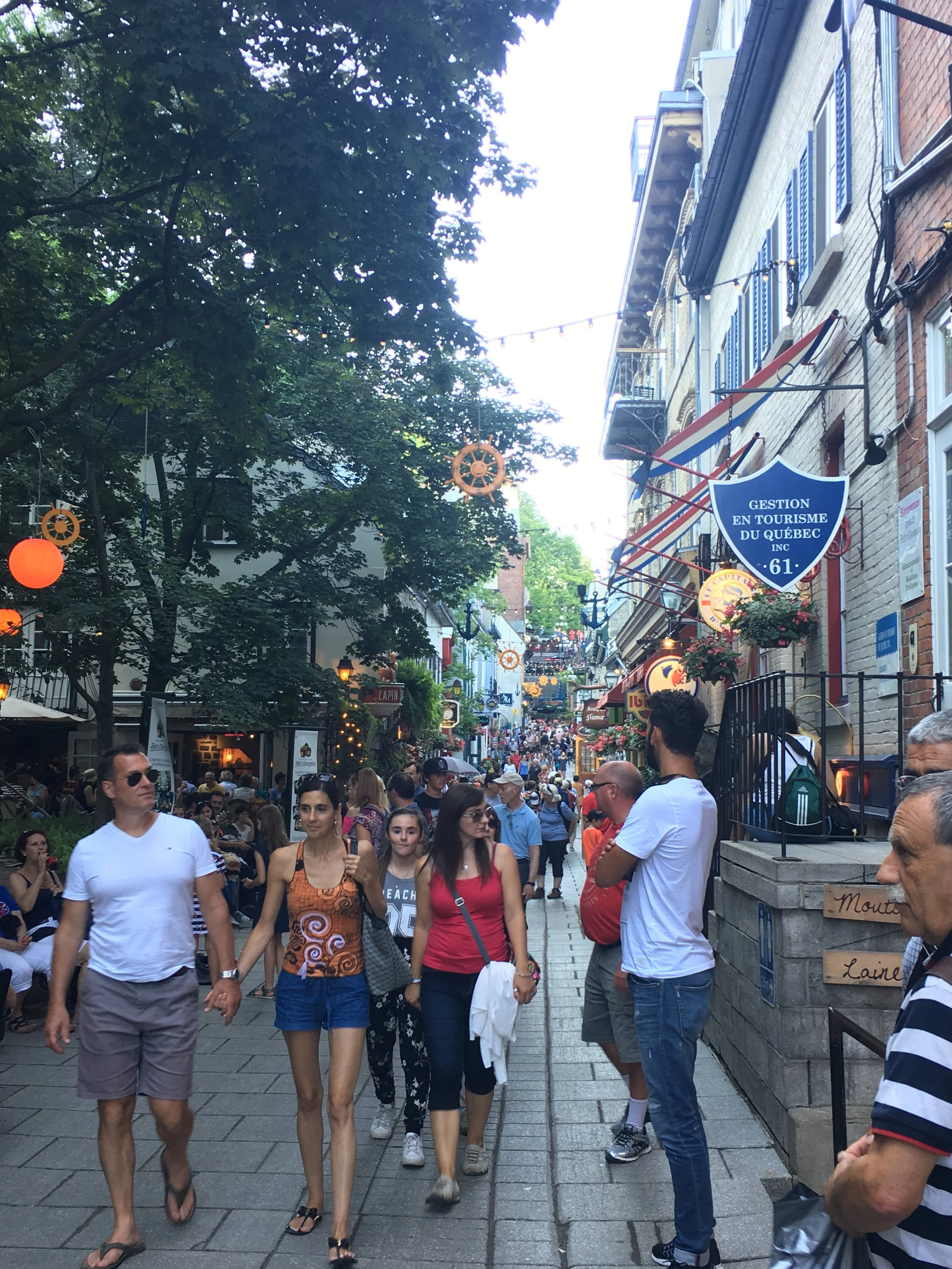 A busy outdoor street scene in Quebec, Canada, with pedestrians walking and sitting at outdoor cafes under decorated string lights. Buildings with balconies line the street, and signs for local businesses are visible.