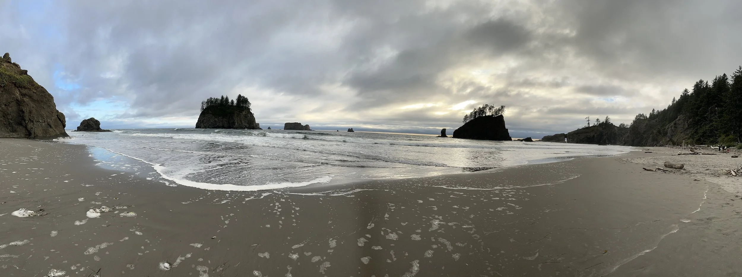 A scenic beach with large rock formations and forested cliffs under a cloudy sky, with waves gently washing onto the sandy shore.