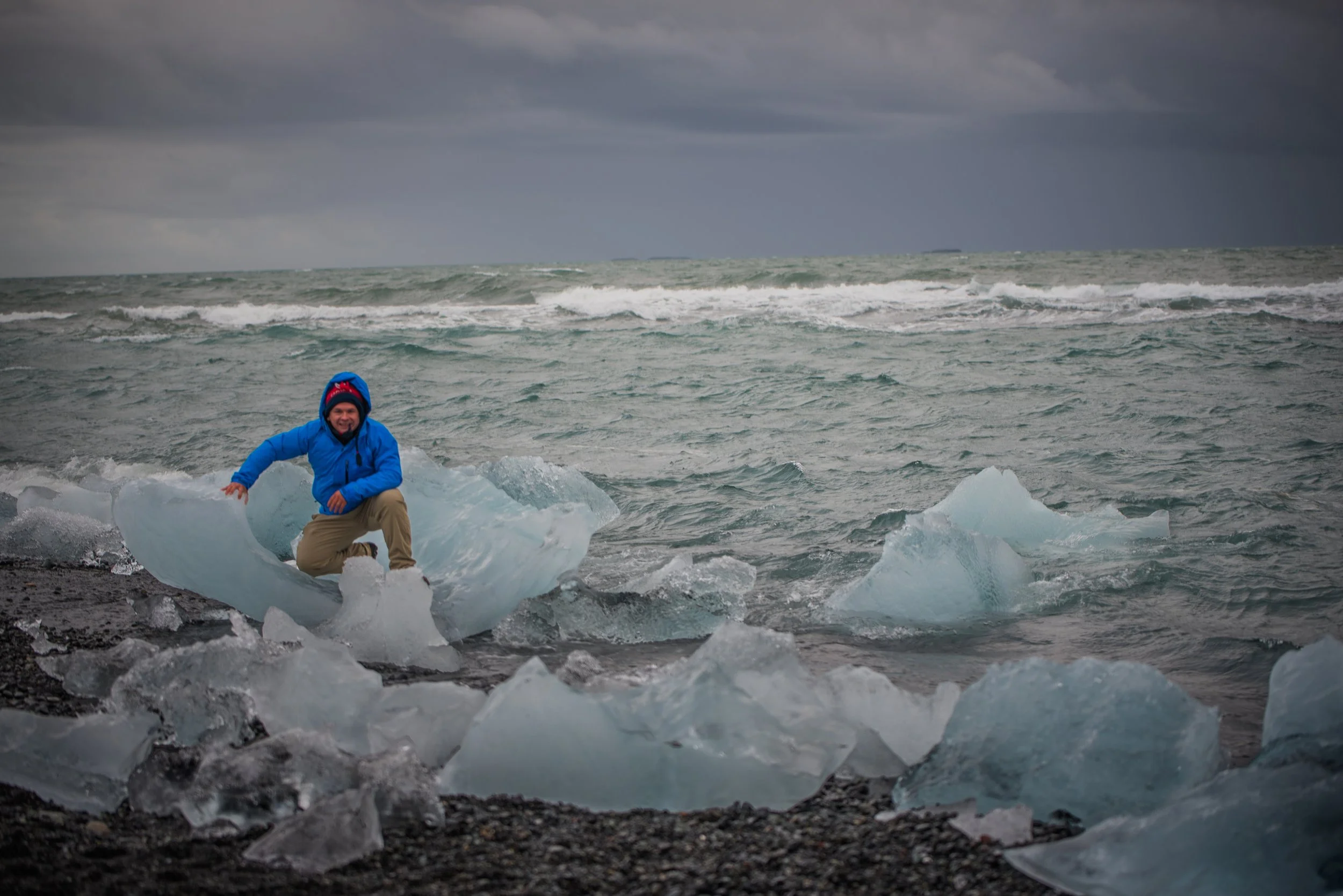 A person in a blue jacket and beige pants kneeling on or near ice chunks along a rocky shoreline, with a rough sea and cloudy sky in the background.
