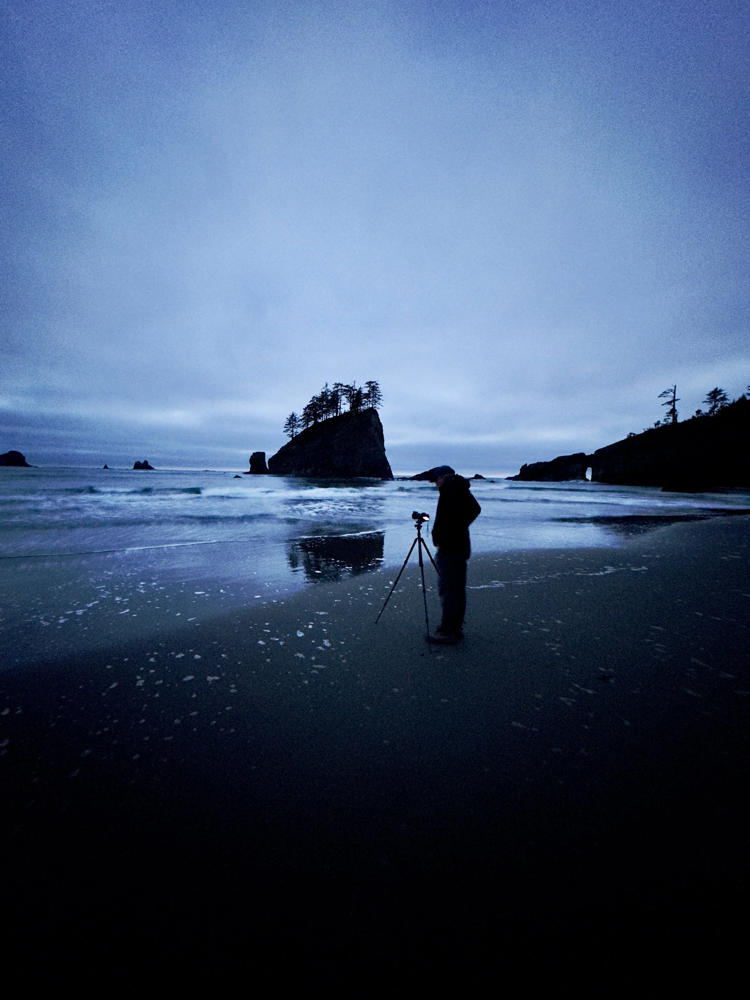 Person standing on a beach at dusk or dawn, setting up a camera on a tripod, with large rocks and island formations in the background and cloudy sky overhead.