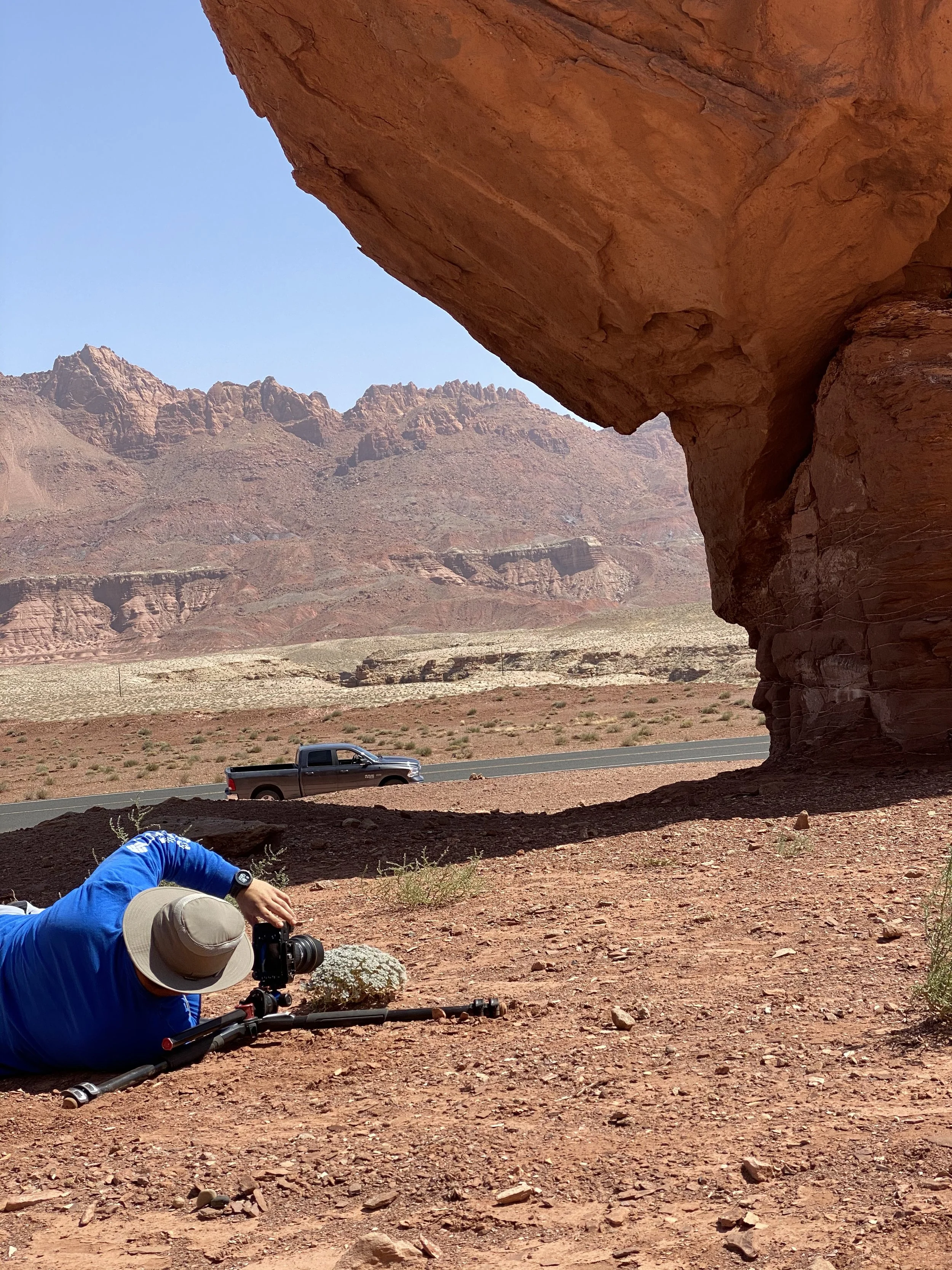 A person lying on the ground with a camera mounted on a tripod, taking photos of a desert landscape with rock formations and mountains in the background, near a large rock overhang.