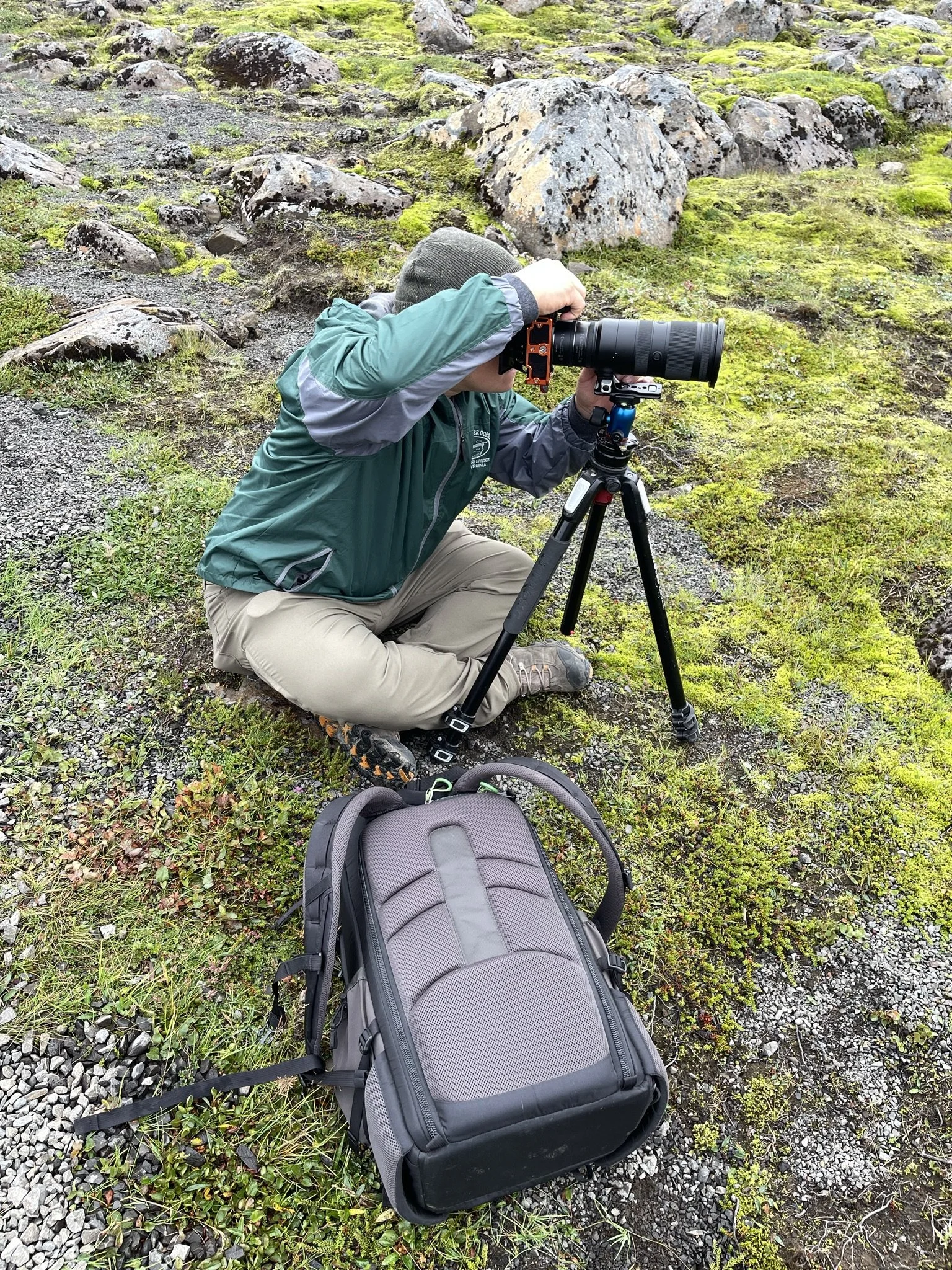 Person kneeling on the ground outdoors, looking through a telescope mounted on a tripod, with a backpack nearby, in a mossy, rocky terrain