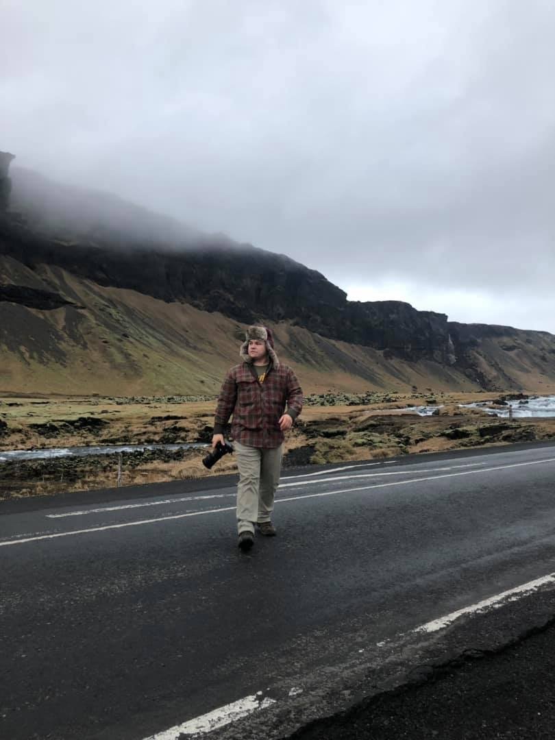Person in a plaid jacket and beige pants walking on a deserted road with cloudy sky and mountains in the background