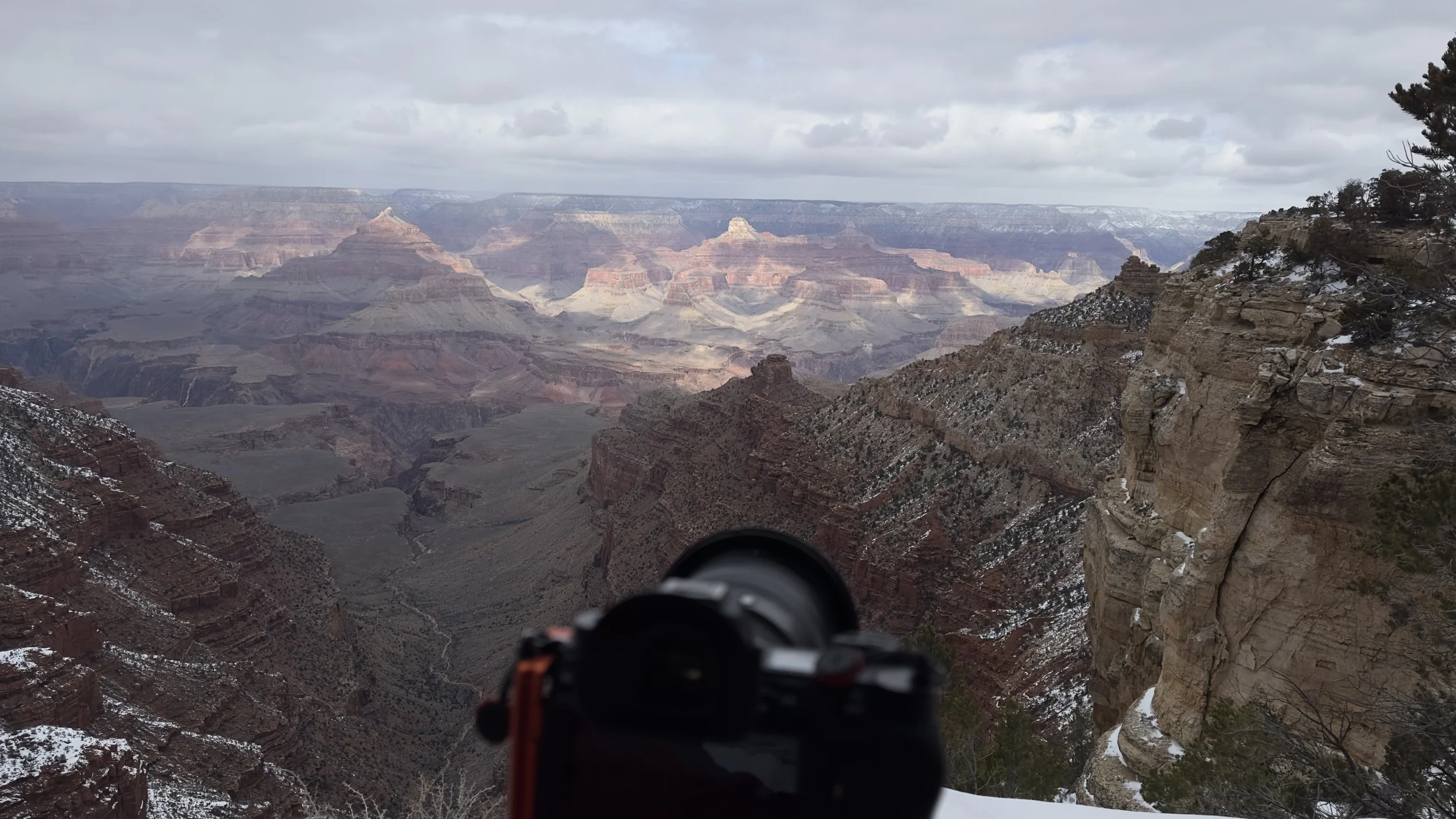 View of the Grand Canyon with a camera in the foreground, showing layered rock formations and a cloudy sky, with patches of snow on the rocky cliffs.