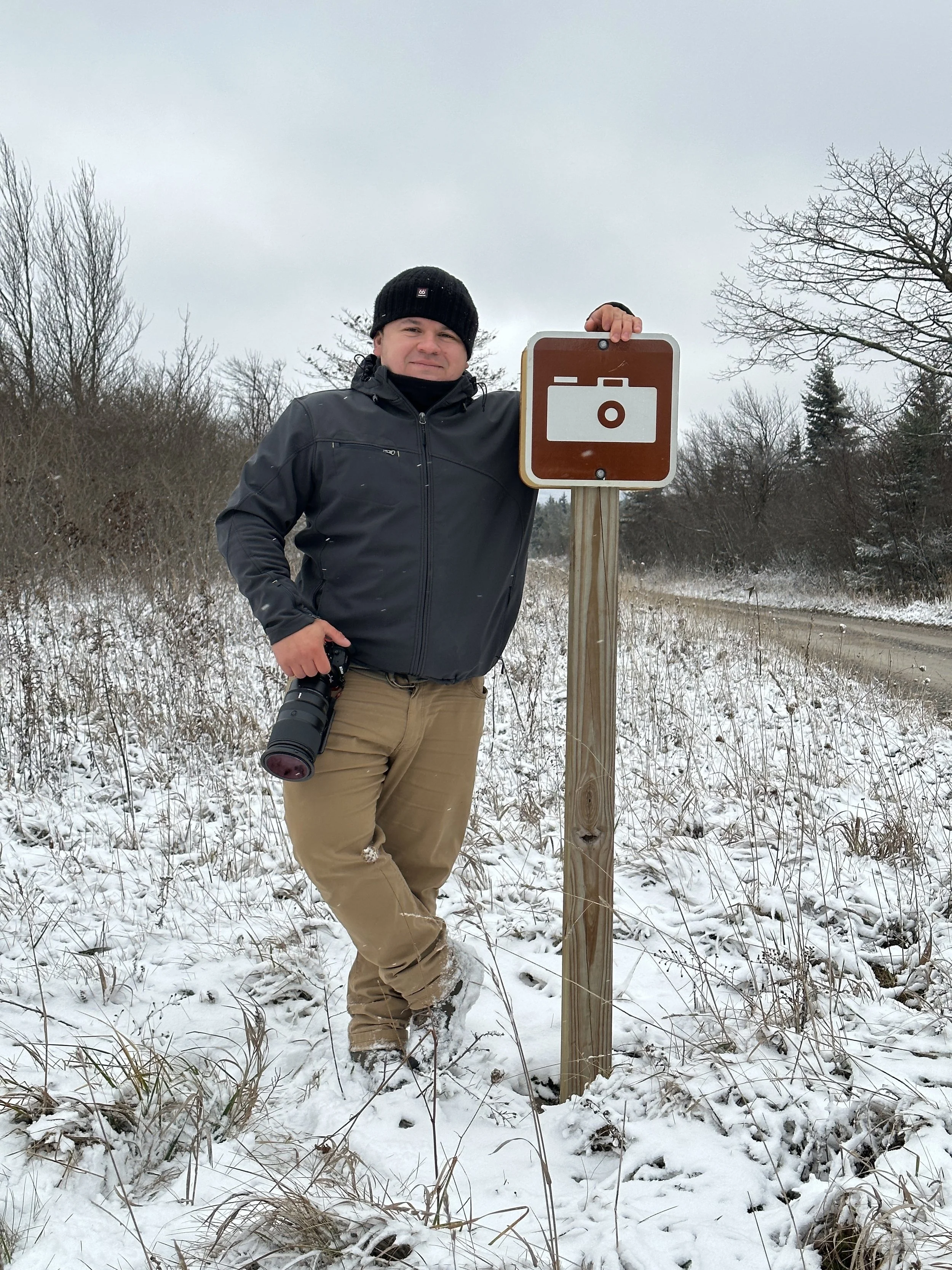 A man standing in a snowy field next to a brown and white photo icon sign, holding a camera and wearing a black jacket, khaki pants, and a black beanie.