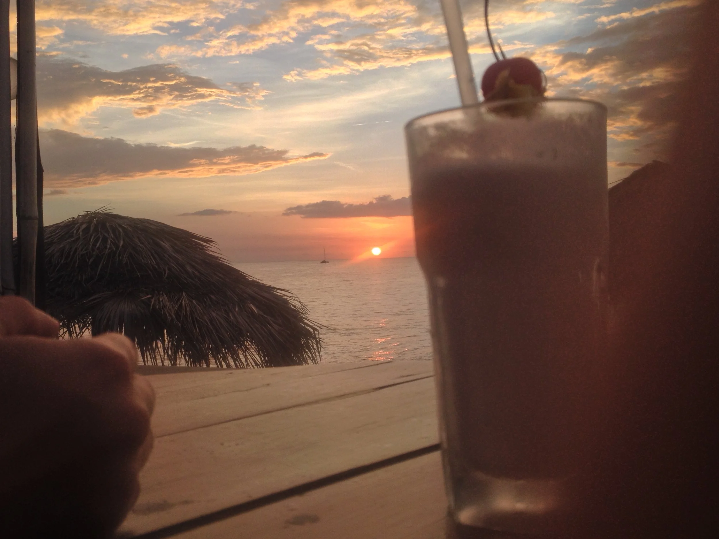 Sunset over the ocean with clouds, a sailboat in the distance, a tiki hut, and a table with a chocolate milkshake topped with a cherry in the foreground.