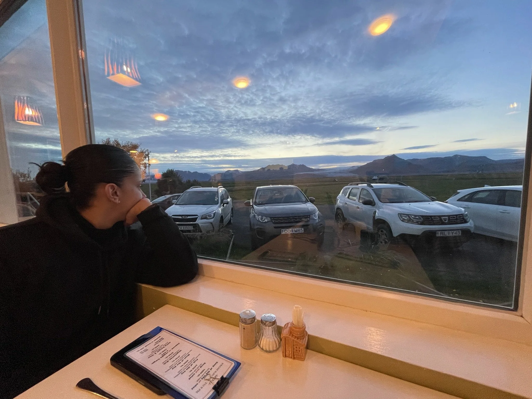A person sitting at a table inside a restaurant, looking out a large window at a sunset or sunrise over a parking lot with several cars and mountains in the distance.
