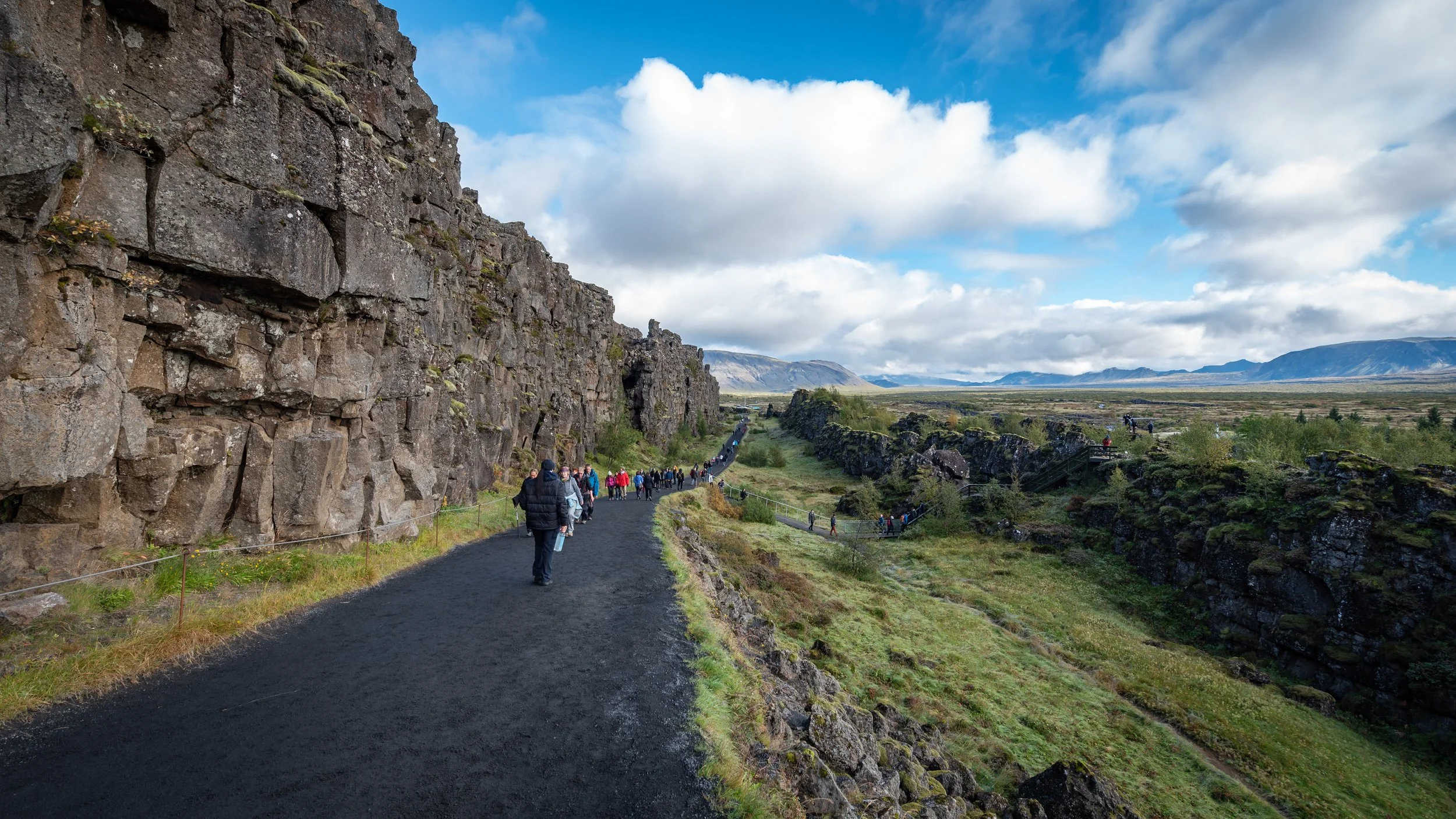 Tourists walking along a mountain trail beside a tall, rugged volcanic rock cliff with a green valley and distant mountains under partly cloudy skies.