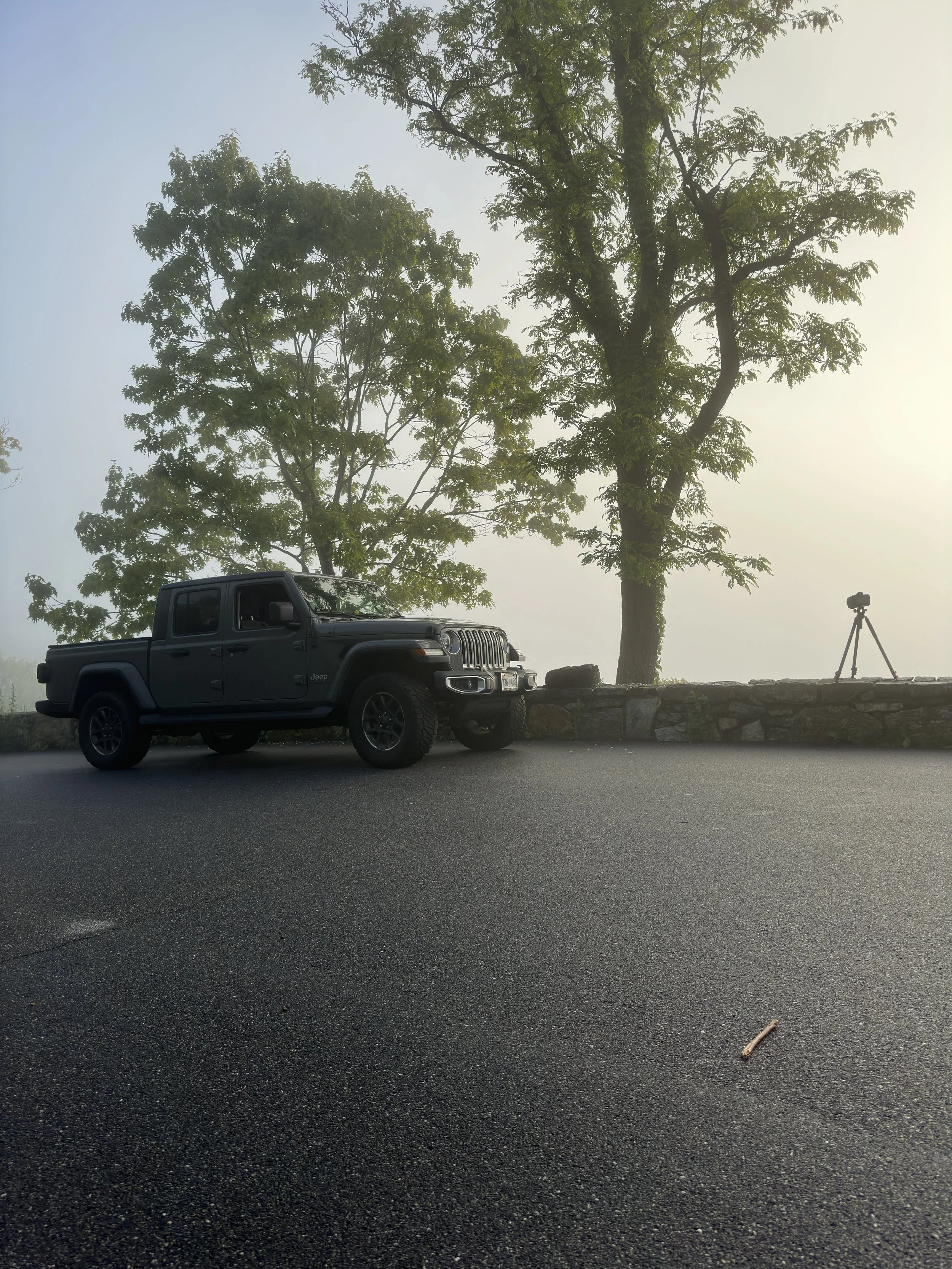 A black Jeep parked on the side of a mountain road with trees and fog in the background, and a camera on a tripod set up nearby.