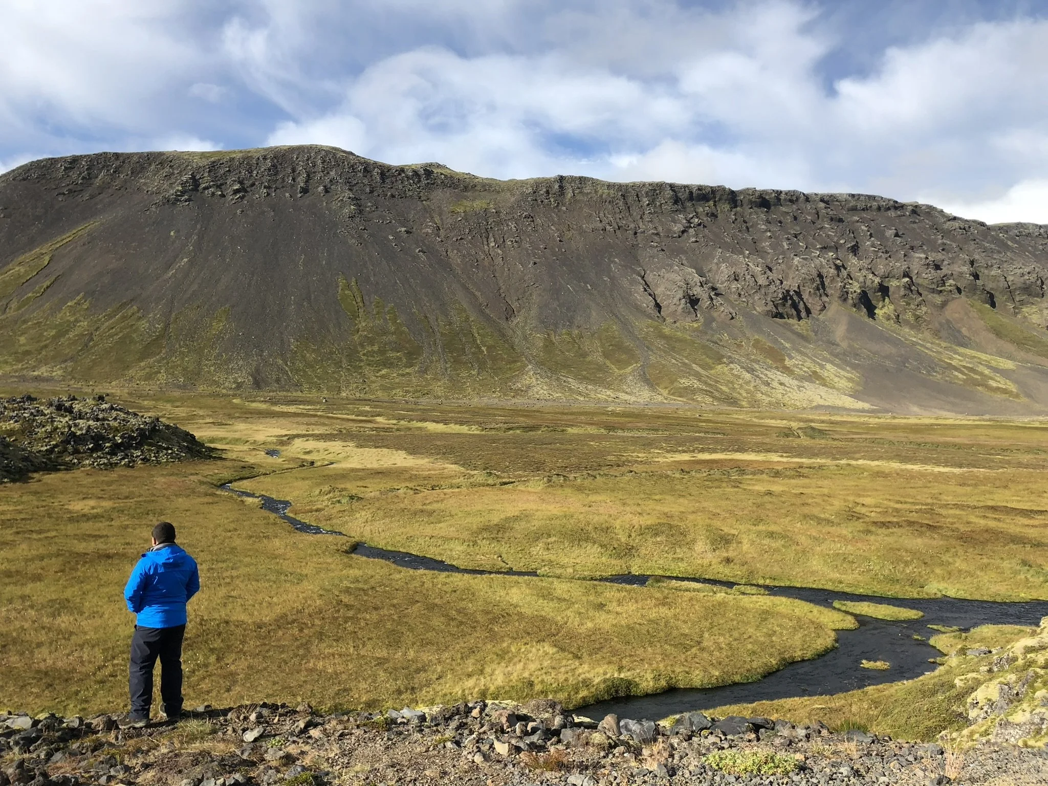 A person in a blue jacket standing on a rocky terrain overlooking a green valley with a winding stream and a large mountain in the background under a partly cloudy sky.