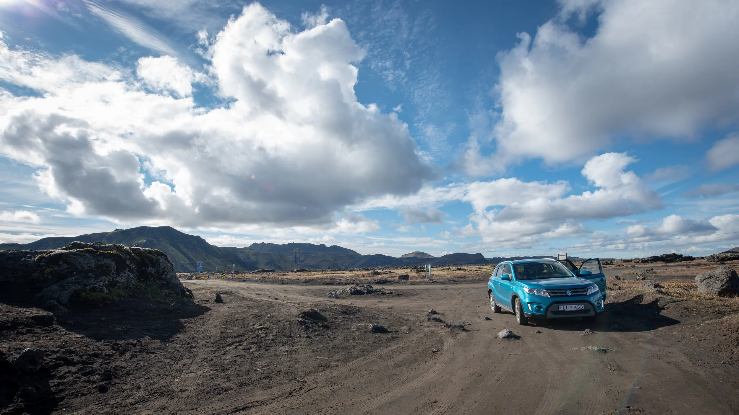A blue Suzuki SUV parked on a dirt road in a rugged landscape with mountains in the background and a partly cloudy sky overhead.