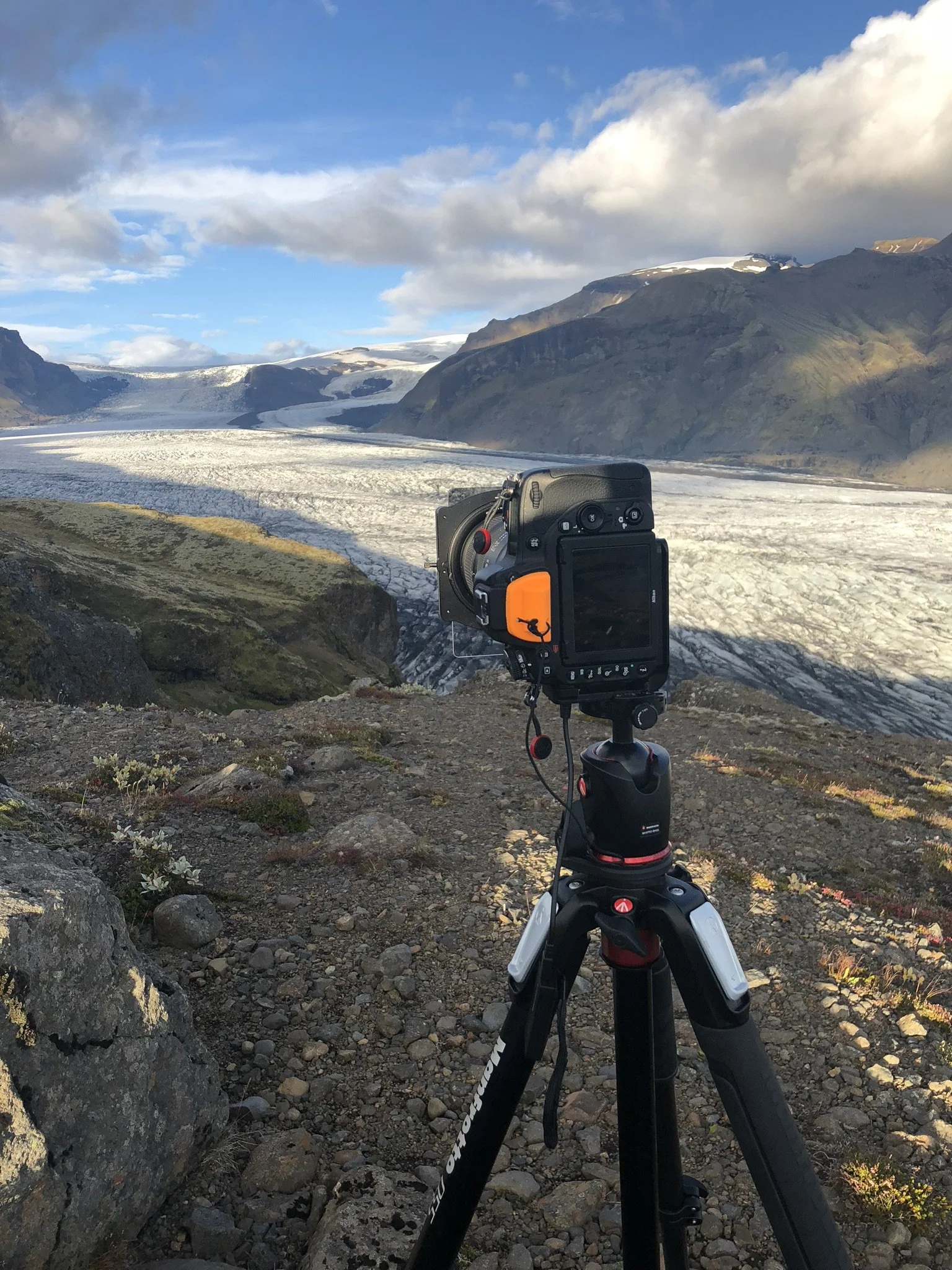 Camera set up on a tripod overlooking a glacier surrounded by mountains and cloudy sky.