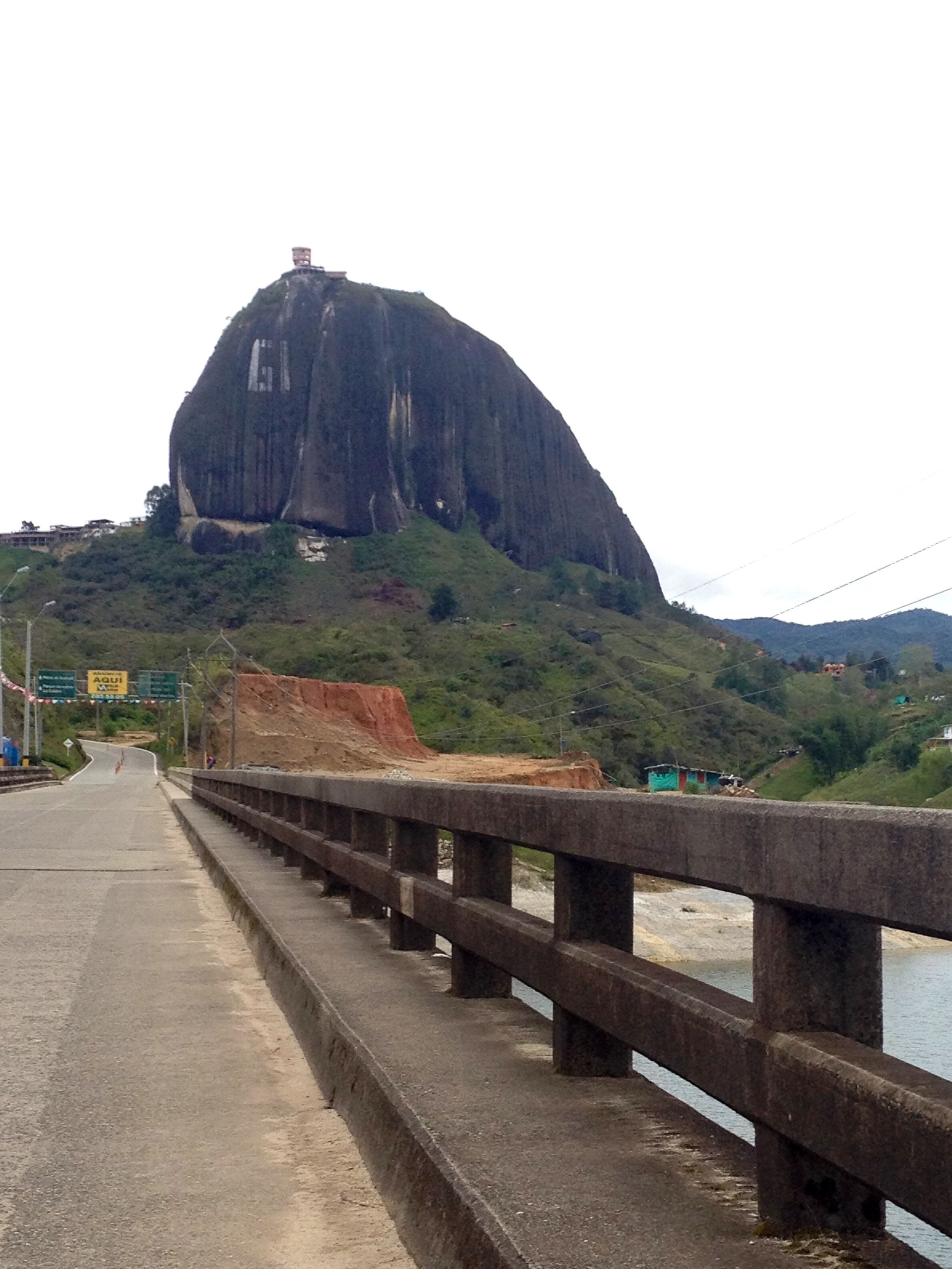 View of Sugarloaf Mountain in Rio de Janeiro, Brazil, with a road and guardrail in the foreground, greenery on the slopes, and cloudy sky.