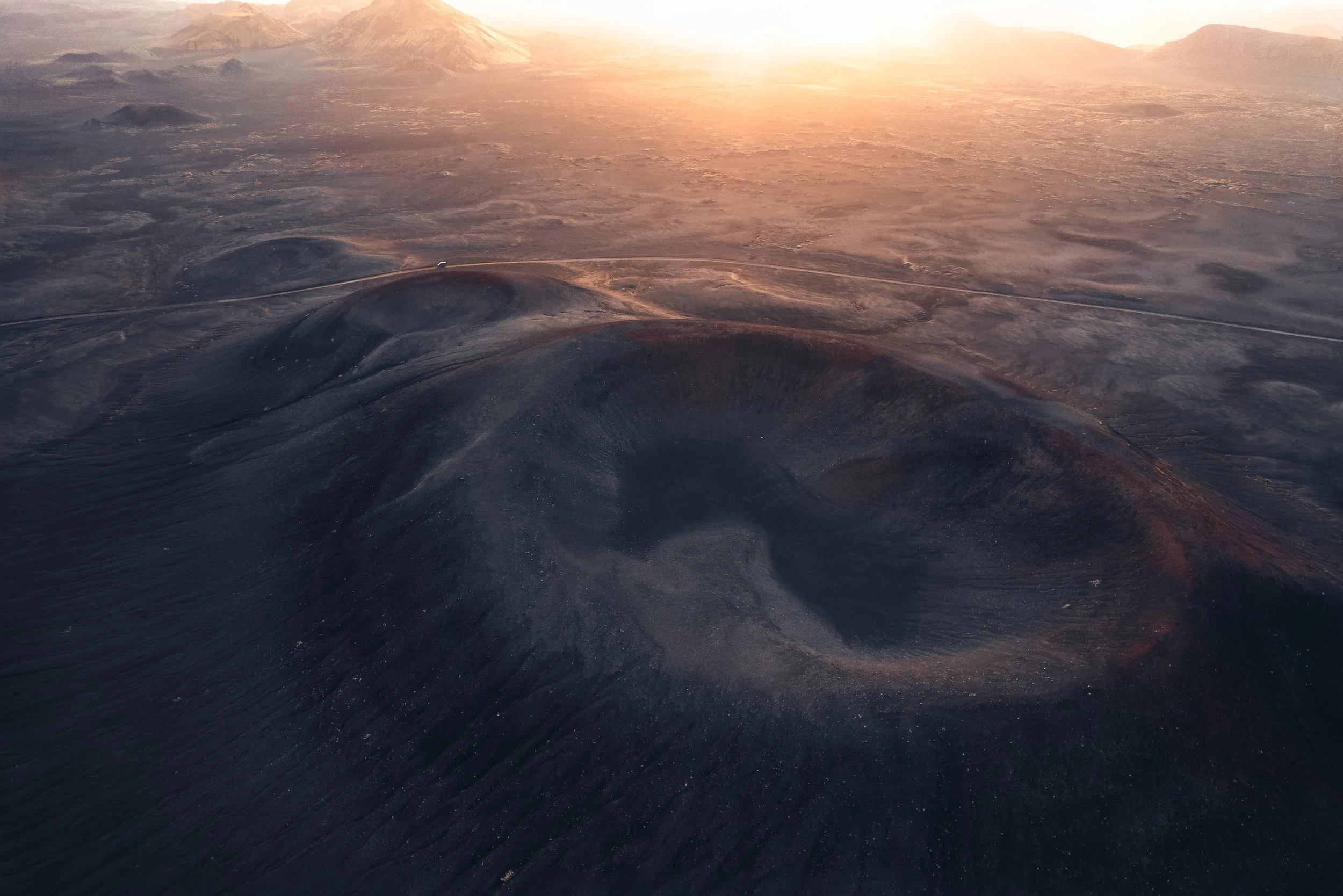 Aerial view of a volcanic crater at sunrise in Iceland's barren landscape, with a solitary vehicle on a winding road.