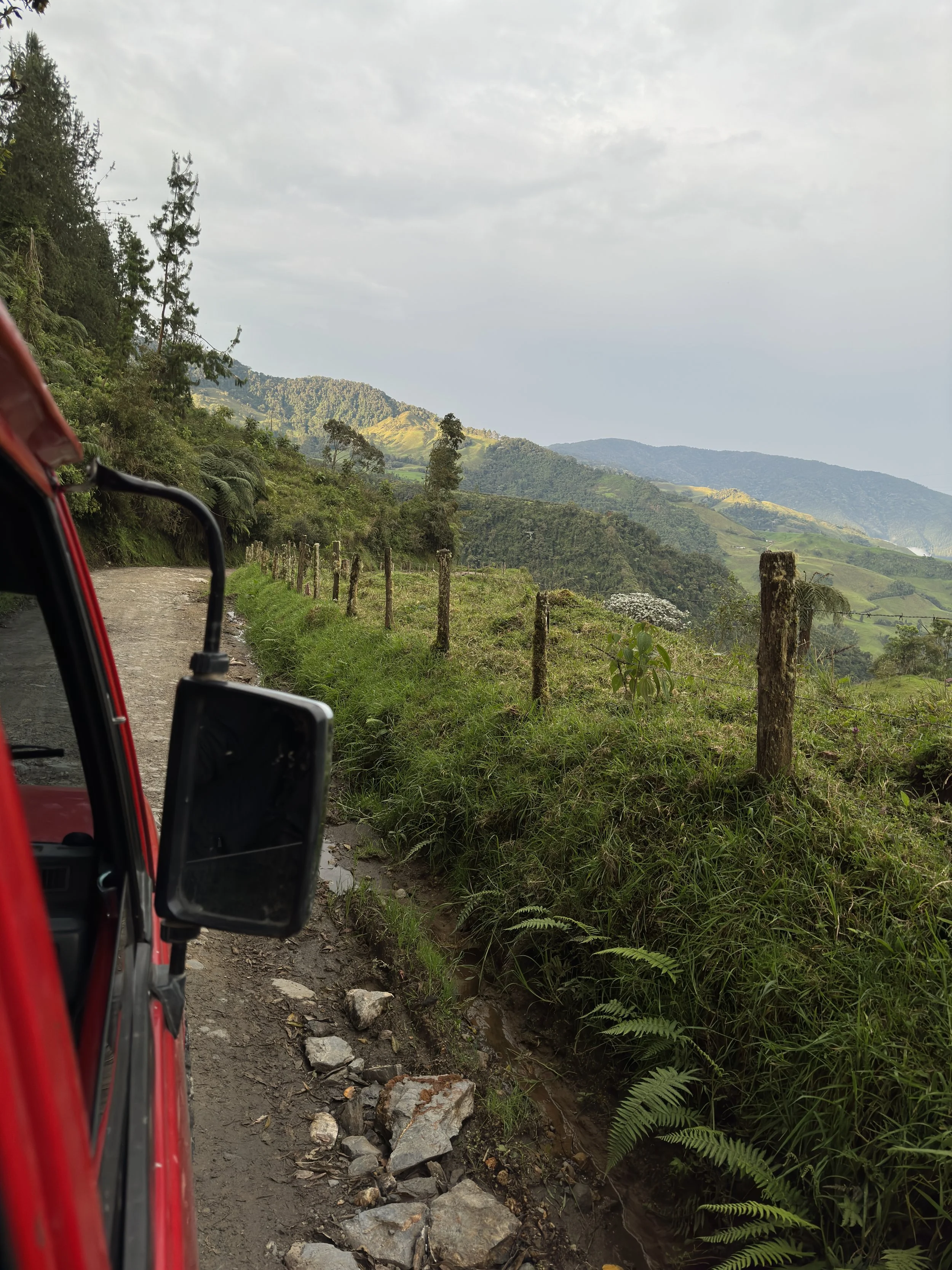 Red vehicle on a dirt road along a green hillside with mountains in the background.