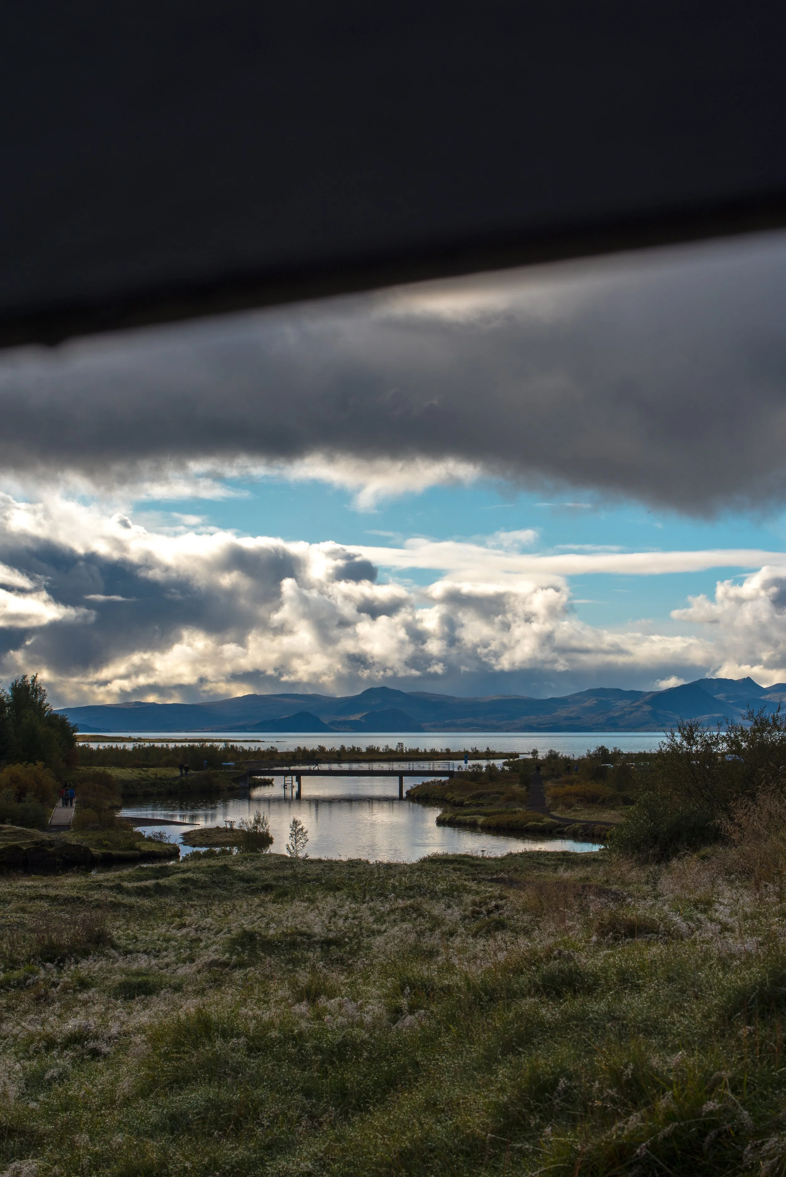 Scenic view of a lake with mountains in the background, cloudy sky, and a bridge over the water, partially obscured by a dark object at the top of the image.