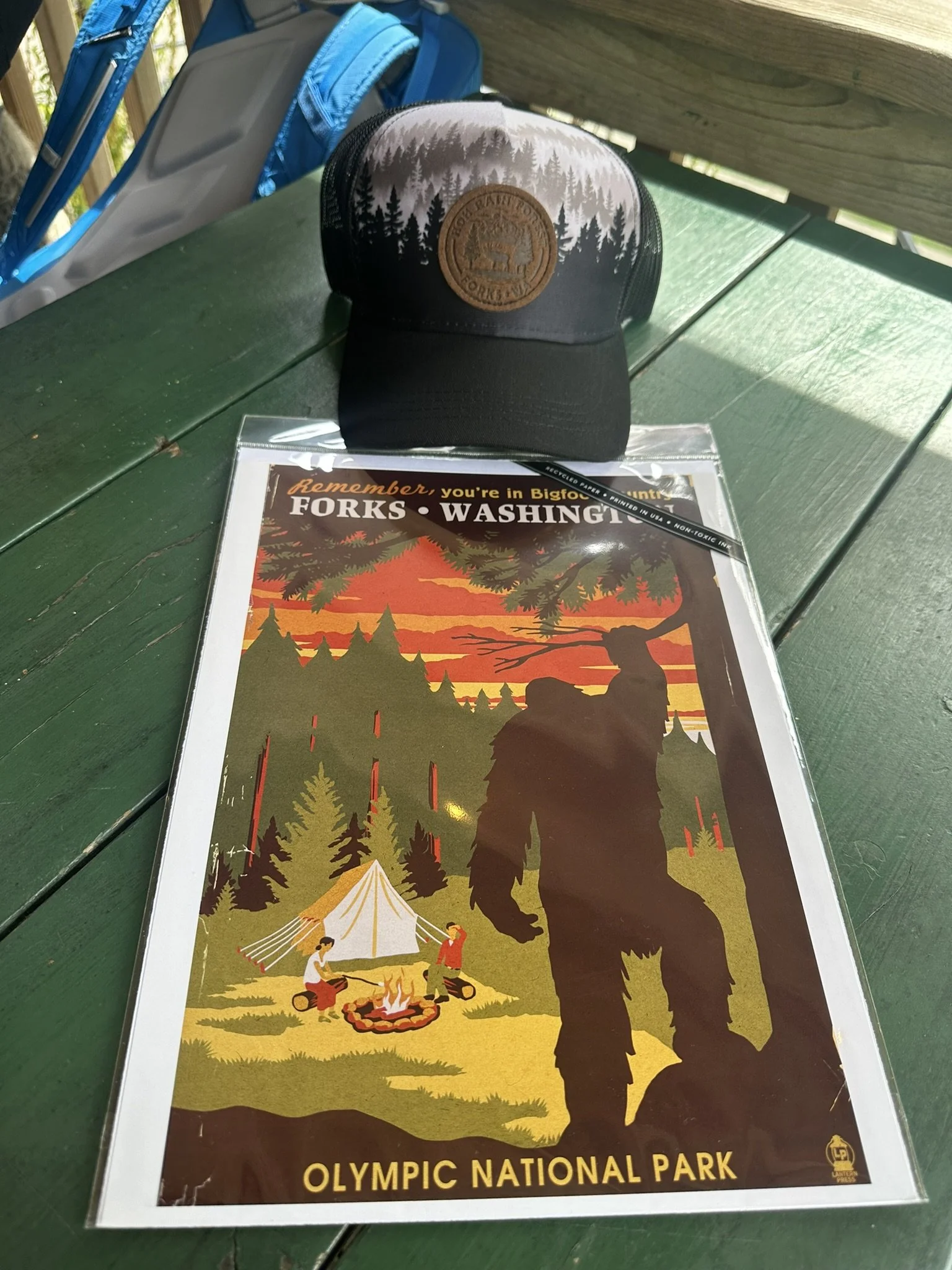 A baseball cap with a forest graphic and leather patch on a green picnic table in Olympic National Park, with a backpack in the background, and a trail map of Forks, Washington, featuring a silhouette of a Bigfoot figure near a camping scene with a t