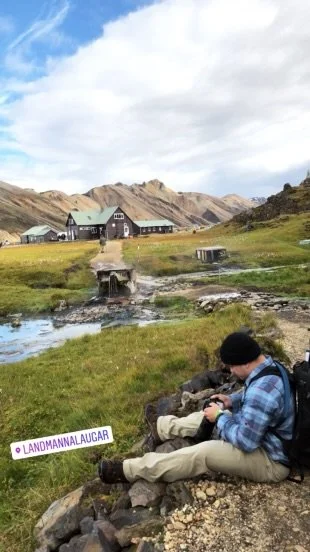 A man sitting on rocks near a stream in a rural landscape with a building in the background and mountains under a partly cloudy sky.