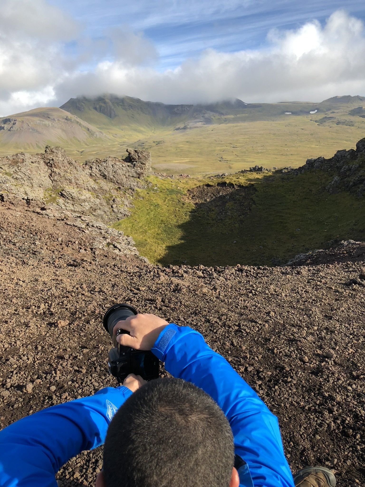 Person in a blue jacket taking a photograph with a camera, sitting on rocky ground with a mountain landscape in the background, including green valleys, rocky formations, and clouds.