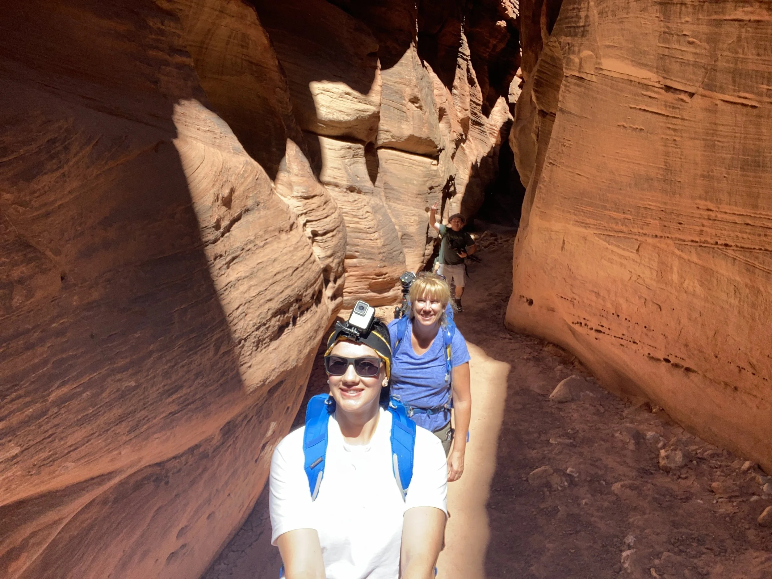 Three women hiking in a narrow slot canyon with red rock walls, one of whom is taking a selfie.
