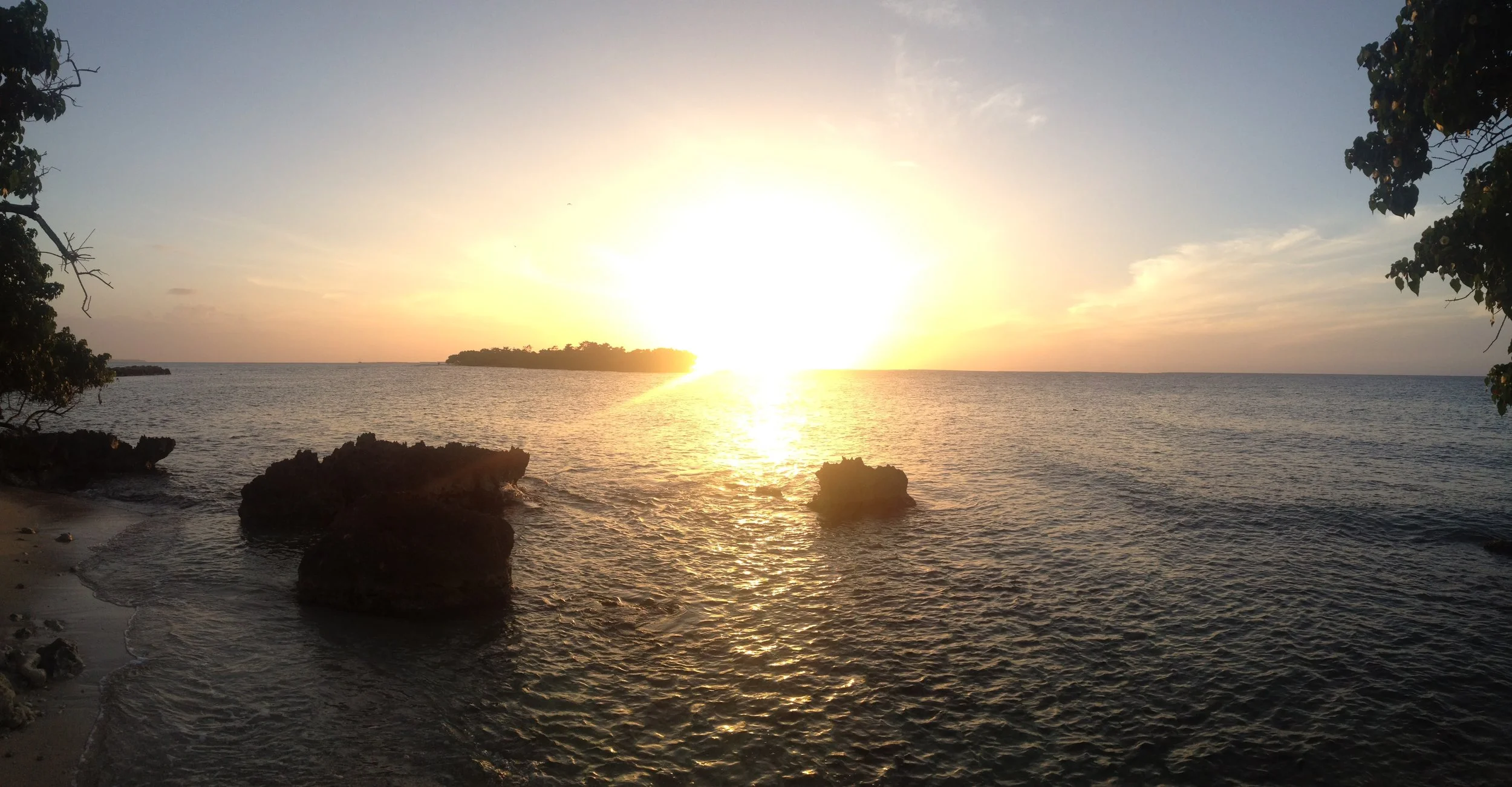 Sunset over the ocean with rocks in the foreground, trees on both sides, and an island in the distance.