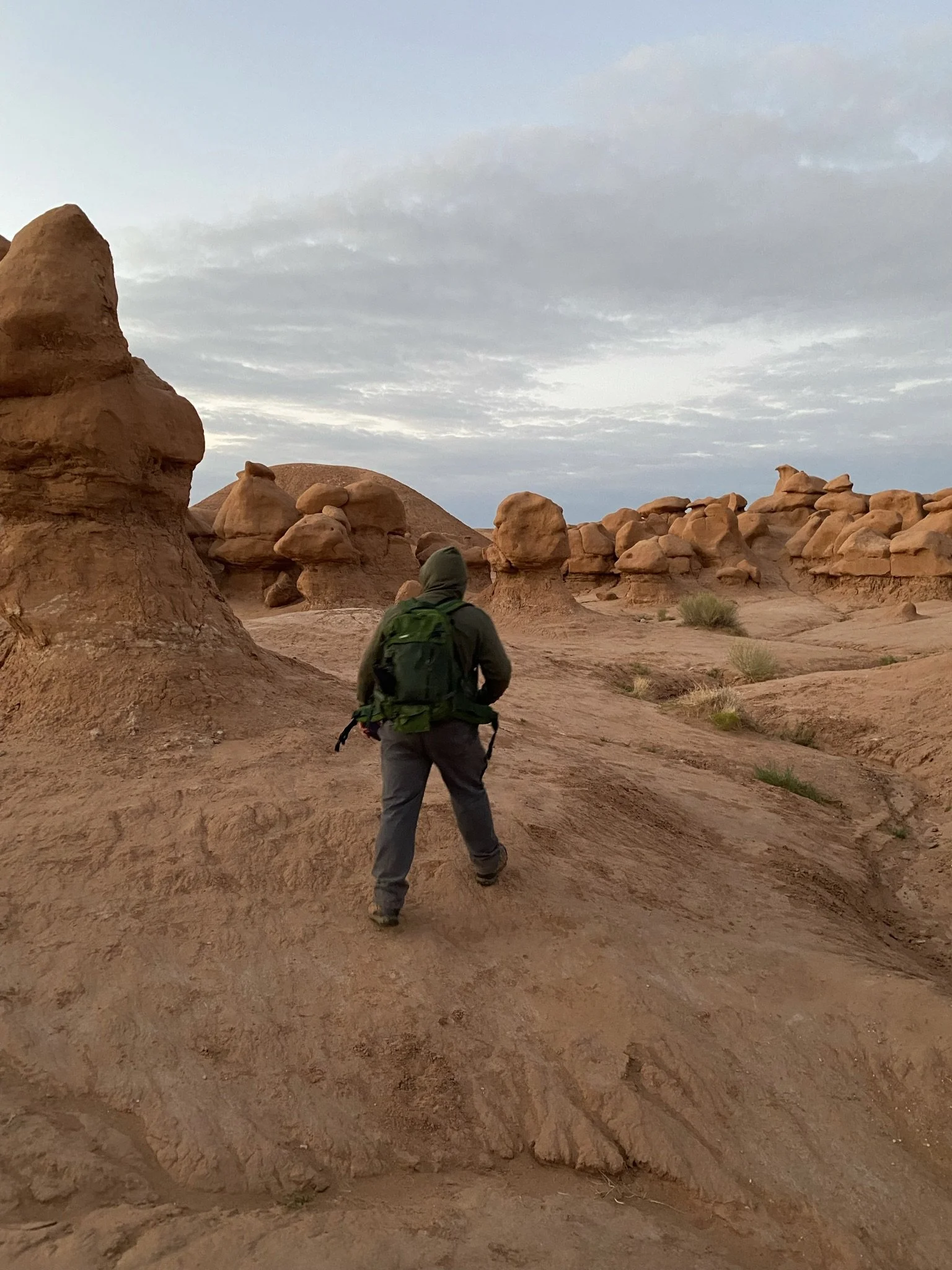 A person with a green backpack walking through a desert with large rock formations and a cloudy sky.
