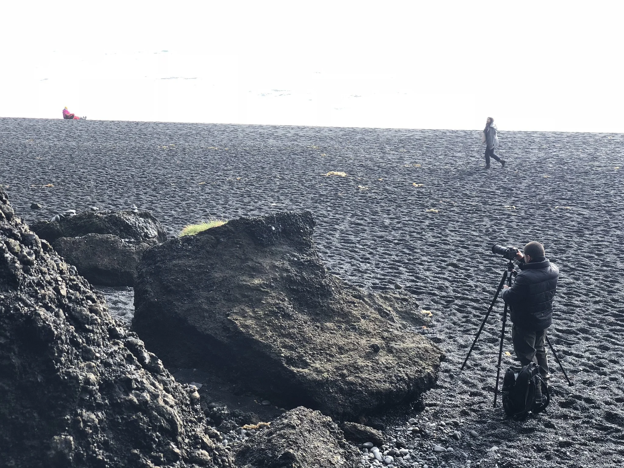 Photographer taking a picture of a woman walking on a black sand beach with rocks, with a person sitting on a pink chair in the distance.
