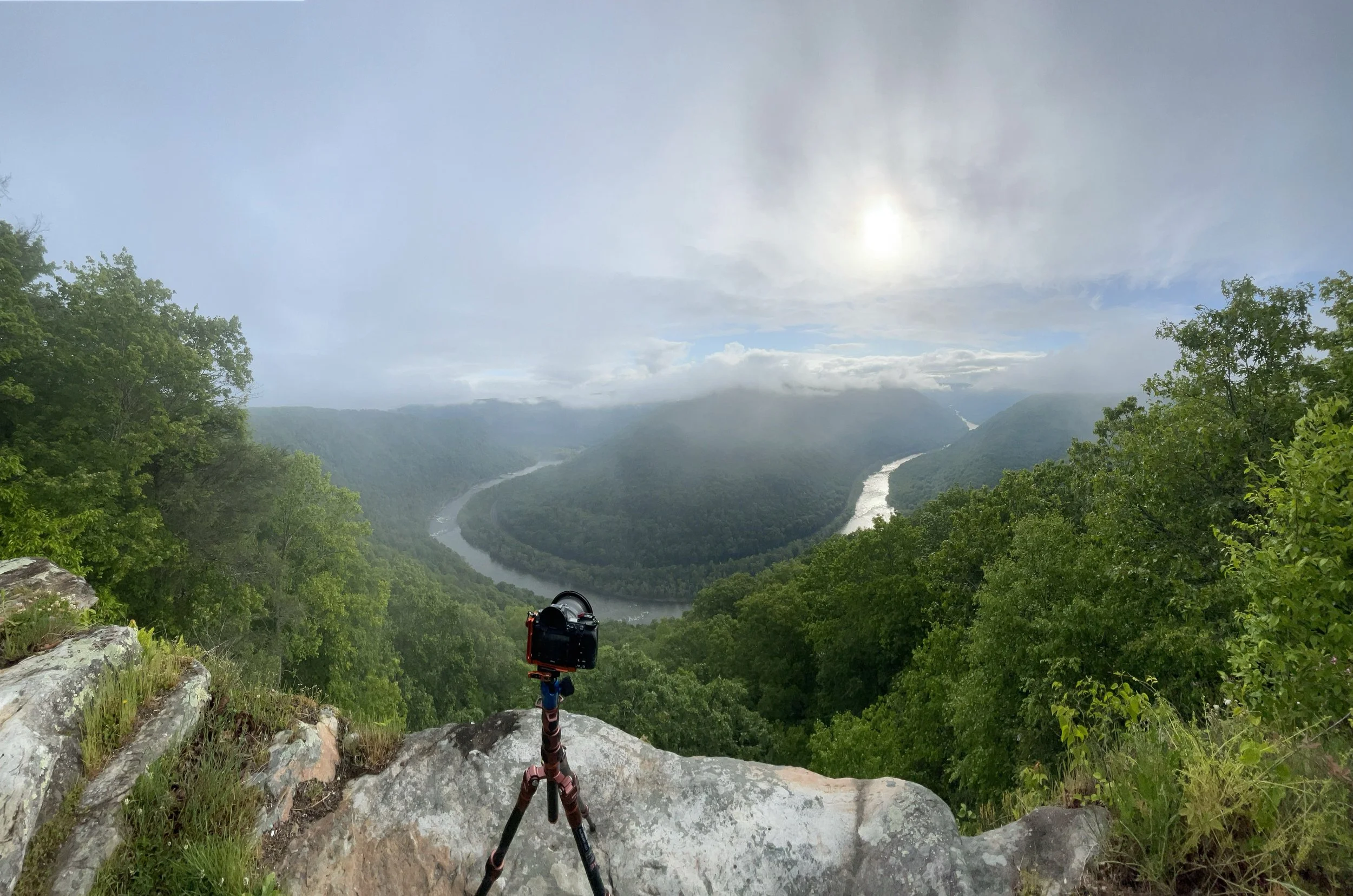 A camera on a tripod positioned on a rocky ledge overlooking a winding river through a lush, green valley with rolling hills, under a partly cloudy sky with the sun peeking through.