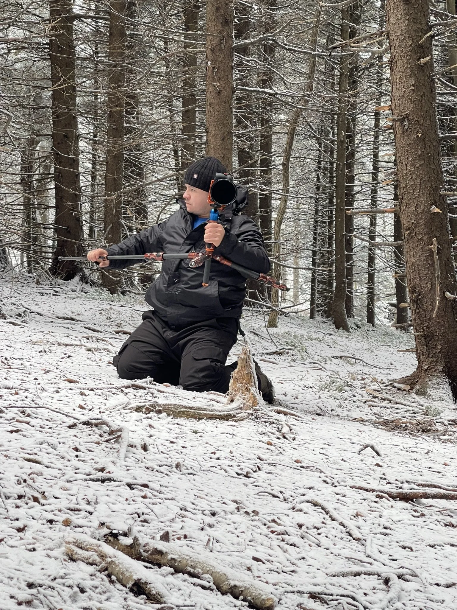 A person kneeling on snow-covered ground in a forest, holding a camera mounted on a tripod, wearing a black jacket, black pants, and a black knit hat.
