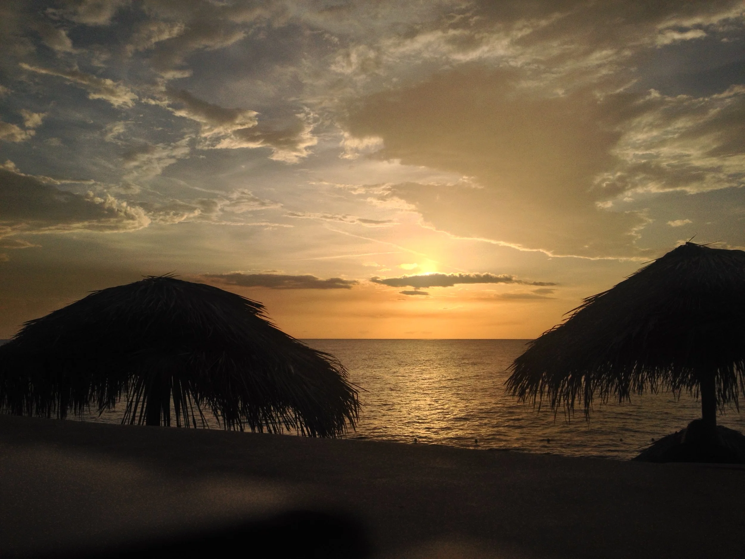 Sunset over the ocean with two thatched umbrellas on the beach in the foreground and clouds in the sky.