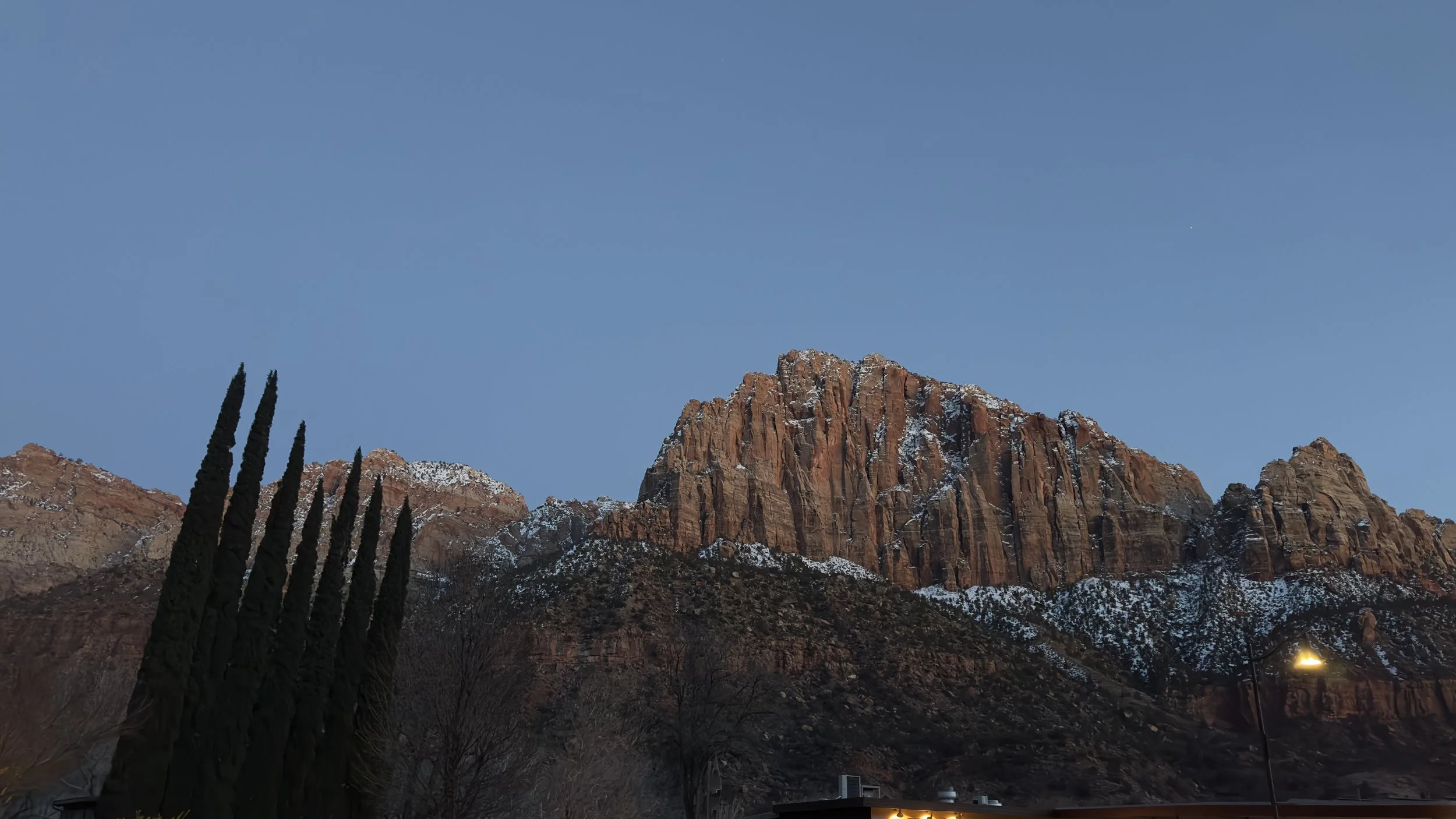 Photograph of rugged mountain with snow patches, dark trees in foreground, and a clear evening sky.