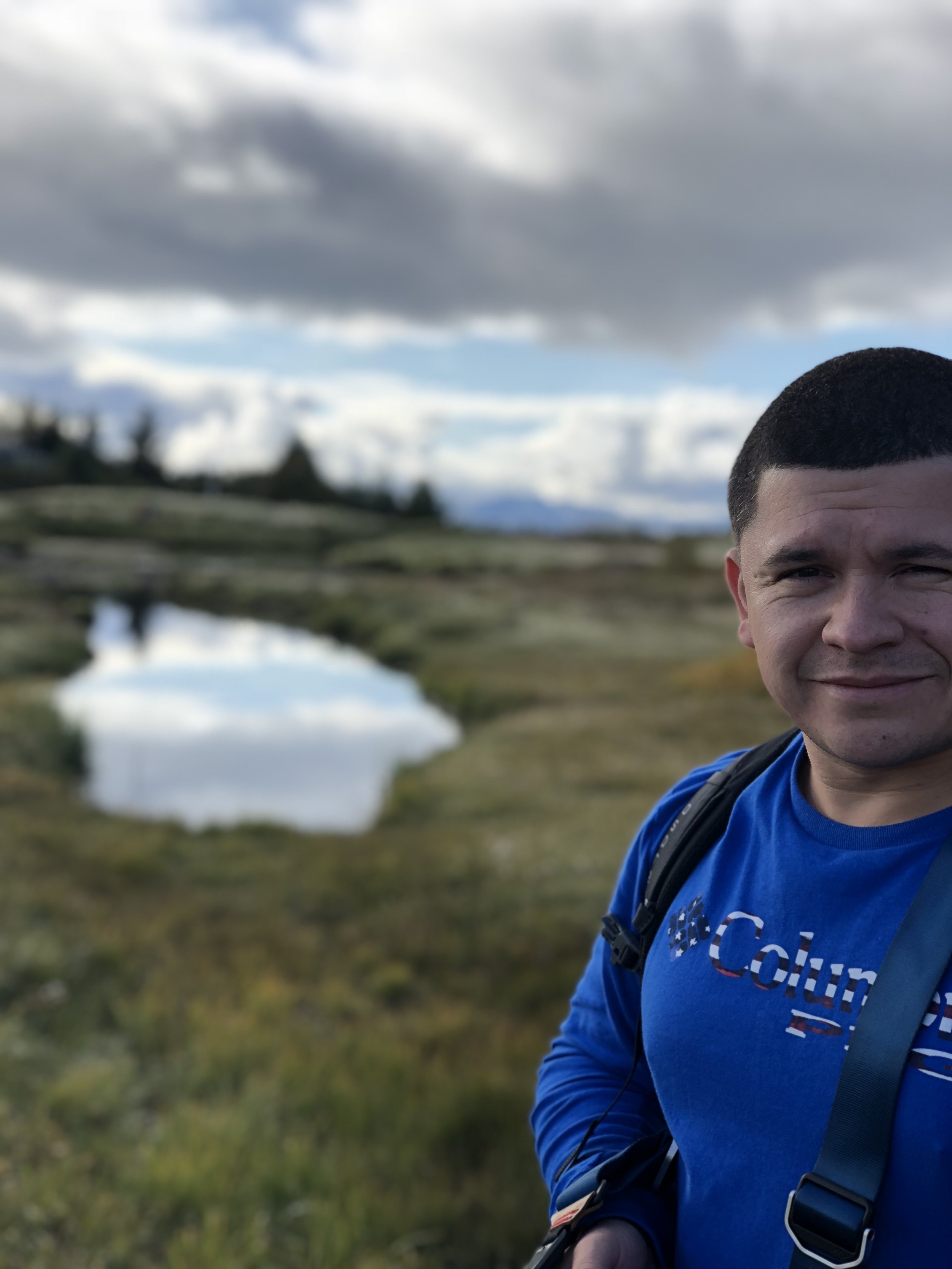 A man with short dark hair wearing a blue long-sleeve shirt and backpack stands outdoors in front of a small lake with reflective water, with cloudy sky and distant trees in the background.
