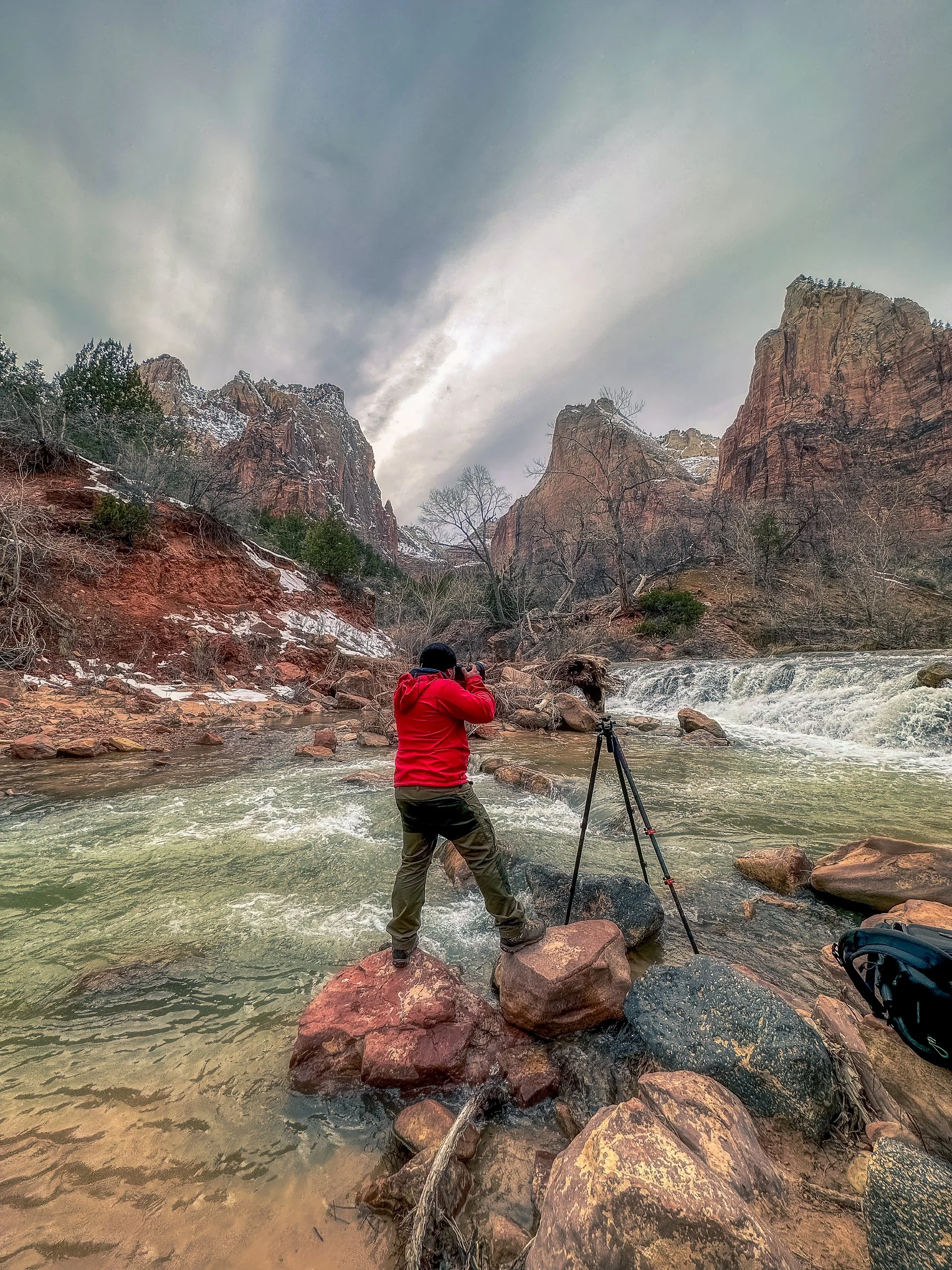 A person in a red jacket taking a photograph with a camera mounted on a tripod while standing on rocks in a river with a waterfall and mountainous landscape in the background.