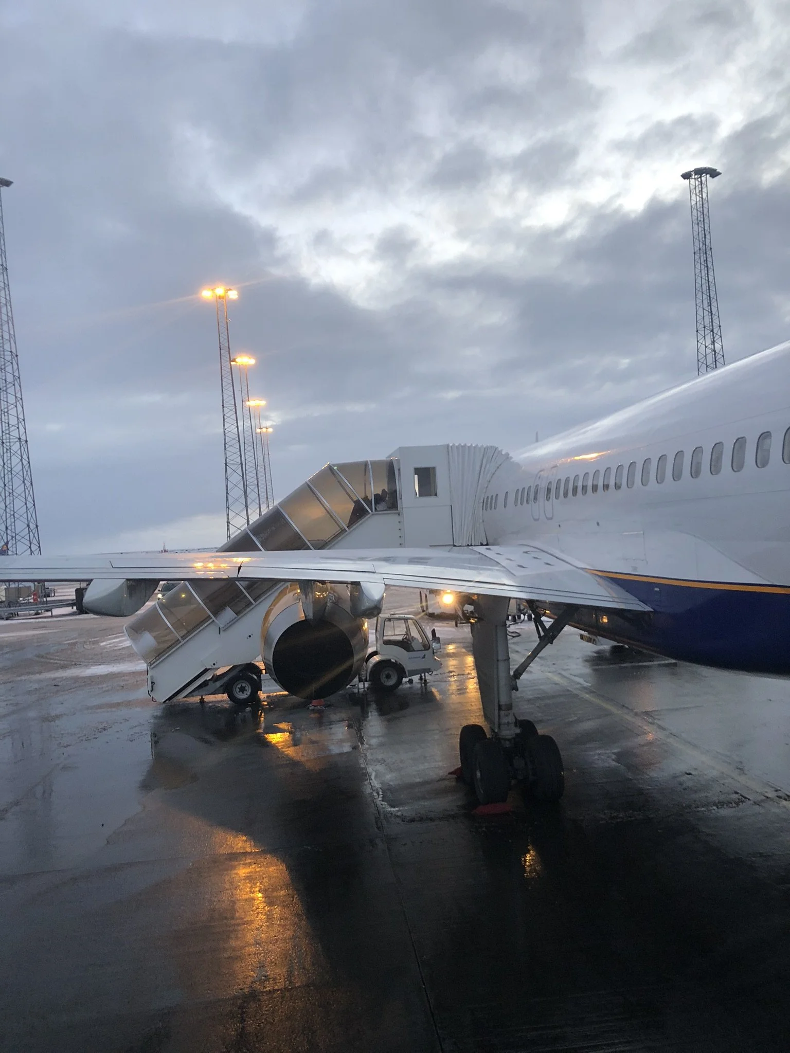 An airplane parked at the airport gate with stairs attached, and a small vehicle underneath. The ground is wet, and the sky is cloudy with the airport lights on.