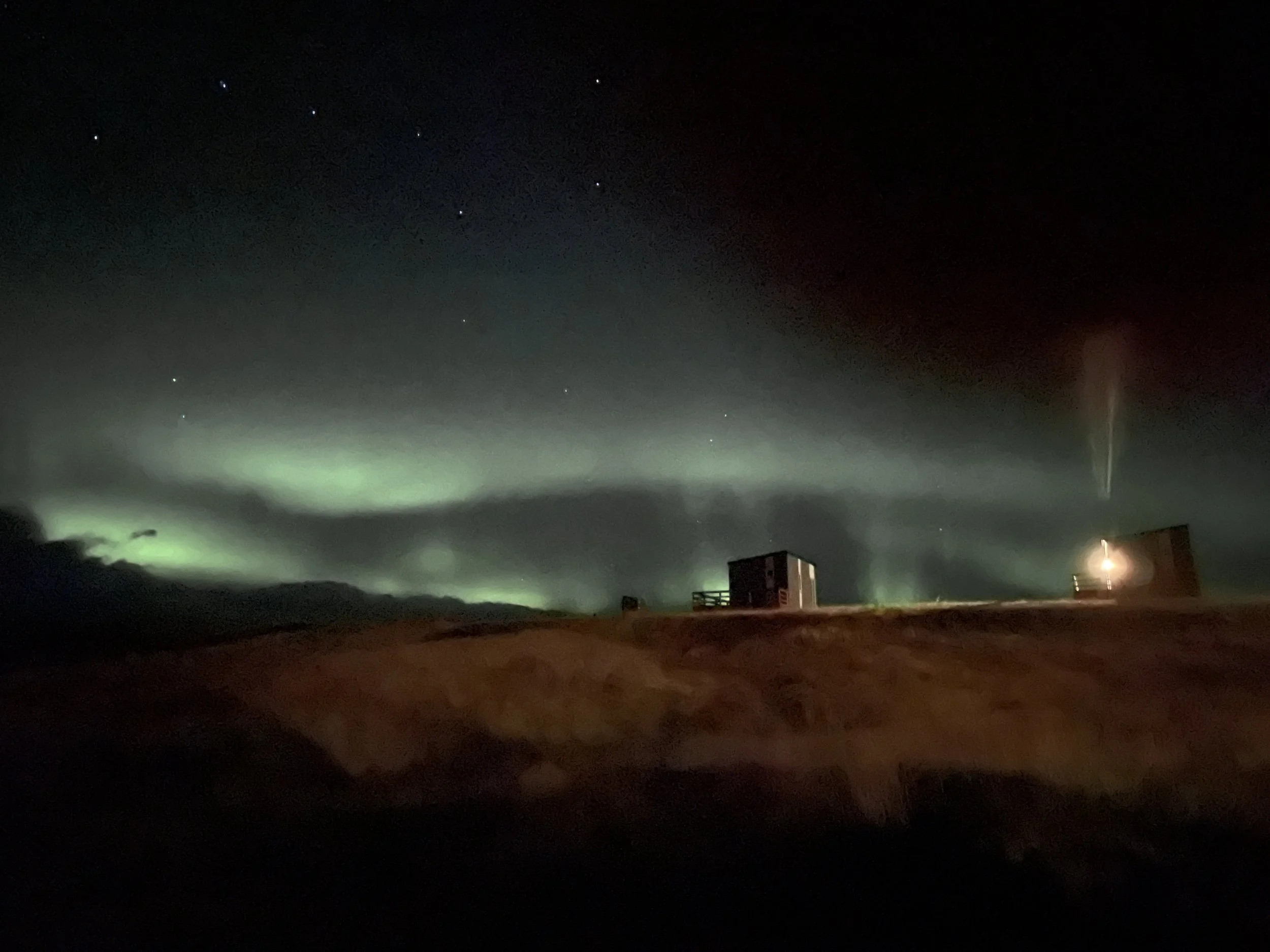 Night sky with northern lights, stars, and a faint rainbow beer-jet of vapor rising from a building, with two small structures on a hill.
