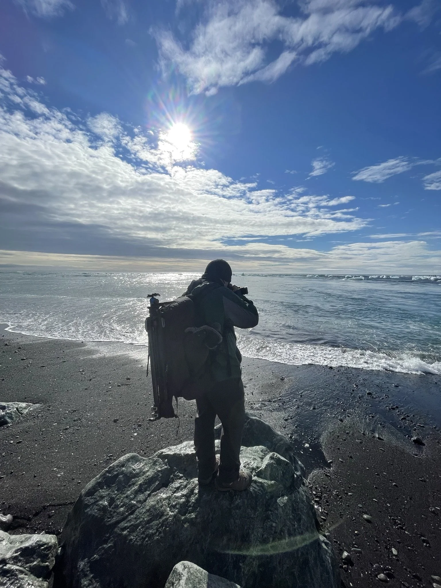 Person standing on a large rock on a rocky beach, taking a photo of the ocean under a partly cloudy sky with the sun shining.