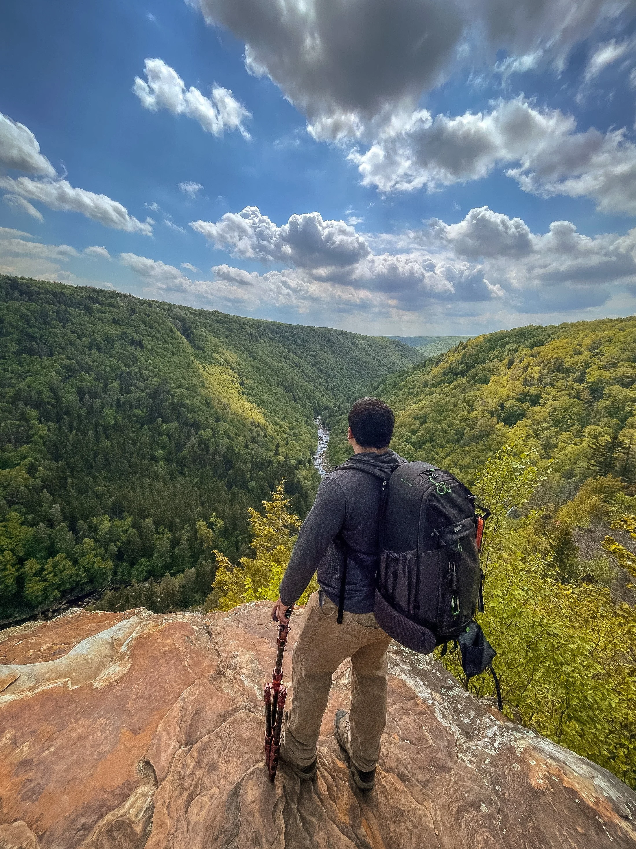 A man standing on a rocky cliff overlooking a green valley and river, carrying a backpack and holding an umbrella, under a cloudy sky.