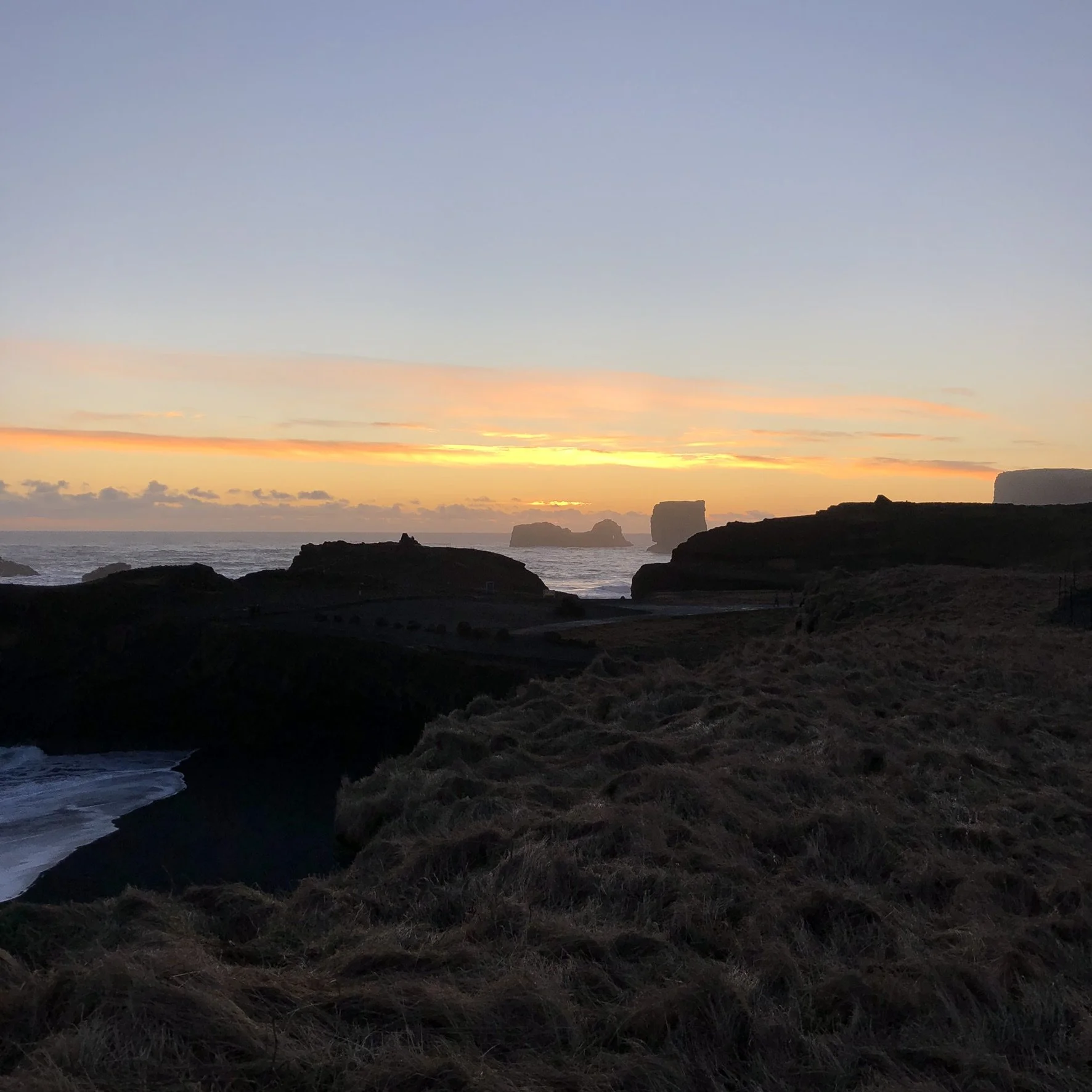A sunset over a rugged coastline with cliffs and rock formations in the distance, with cloudy skies and dark foreground grasses.