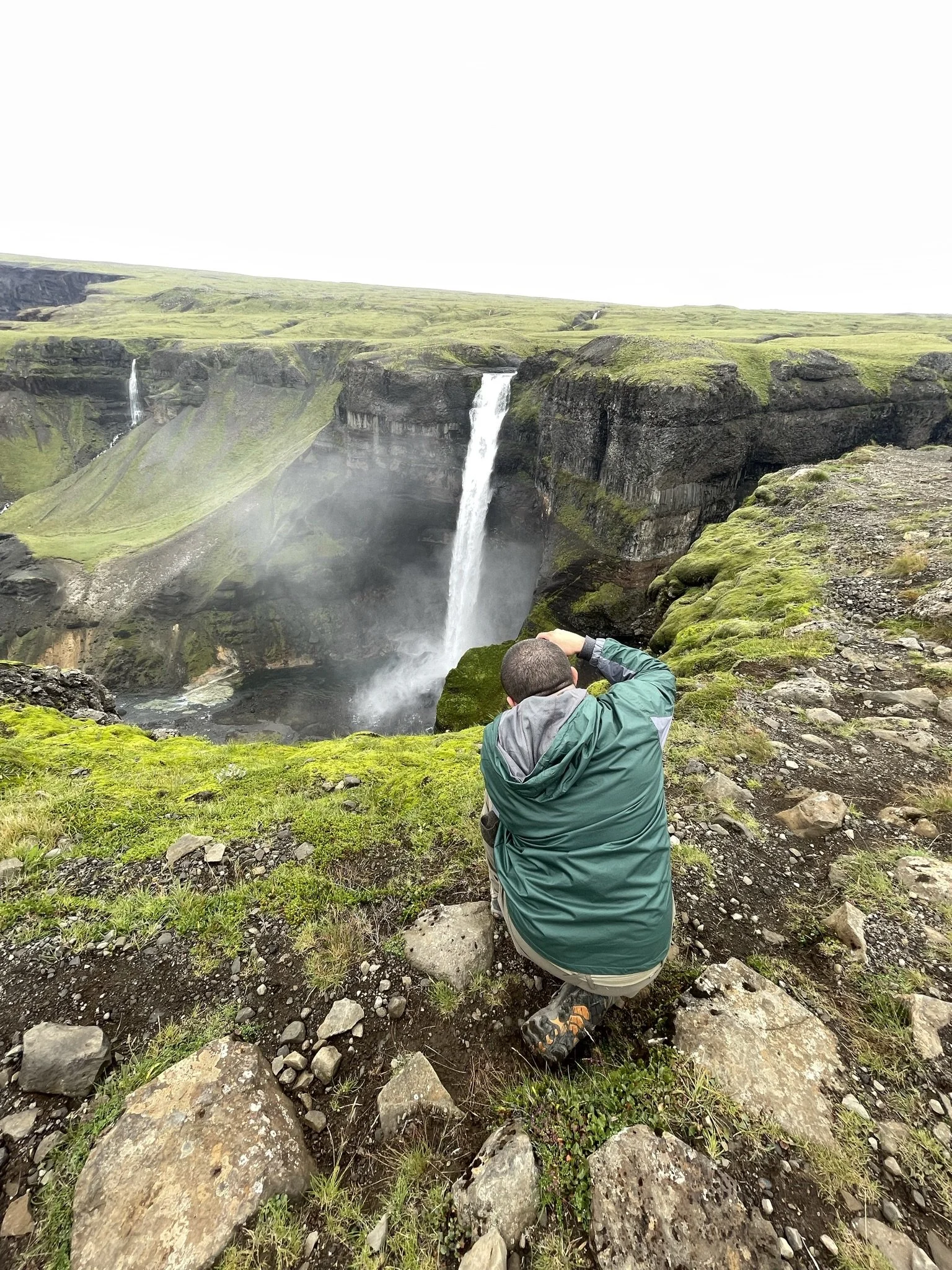 A person in a green jacket kneeling on mossy ground, taking a photograph of a waterfall in a lush green landscape.