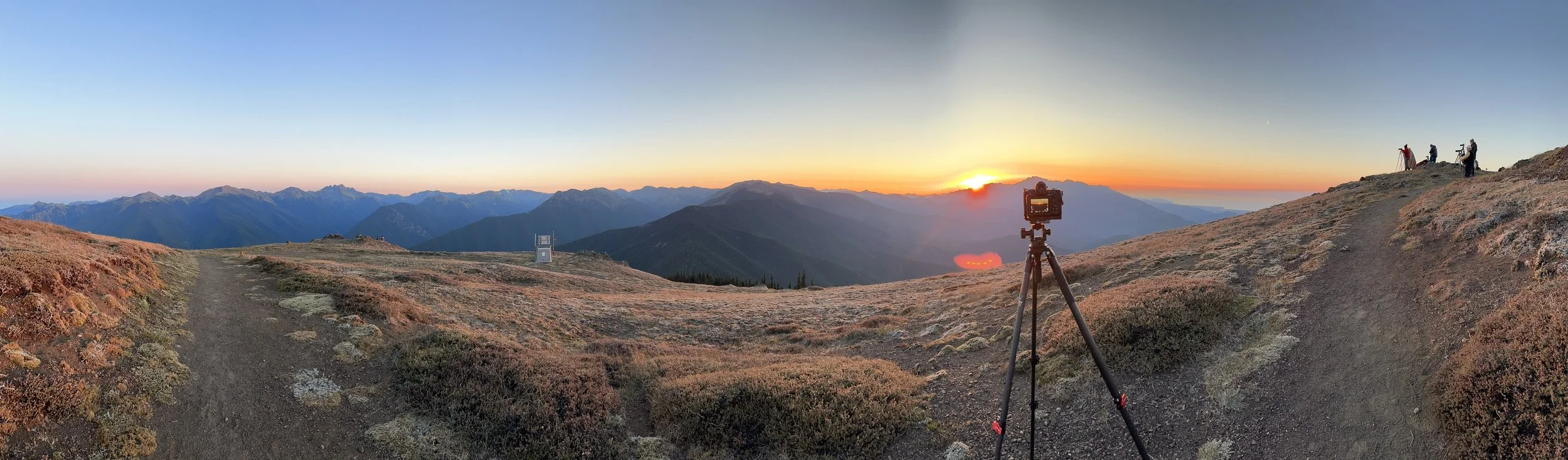 A mountain landscape at sunrise with a camera on a tripod in the foreground, and hikers on a trail to the right.