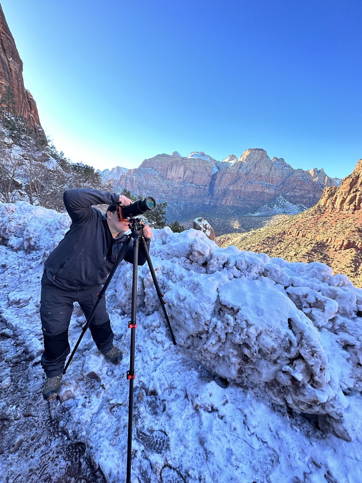 person taking photos with a camera on a tripod in a snowy mountainous landscape with clear blue sky and rocky cliffs in the background