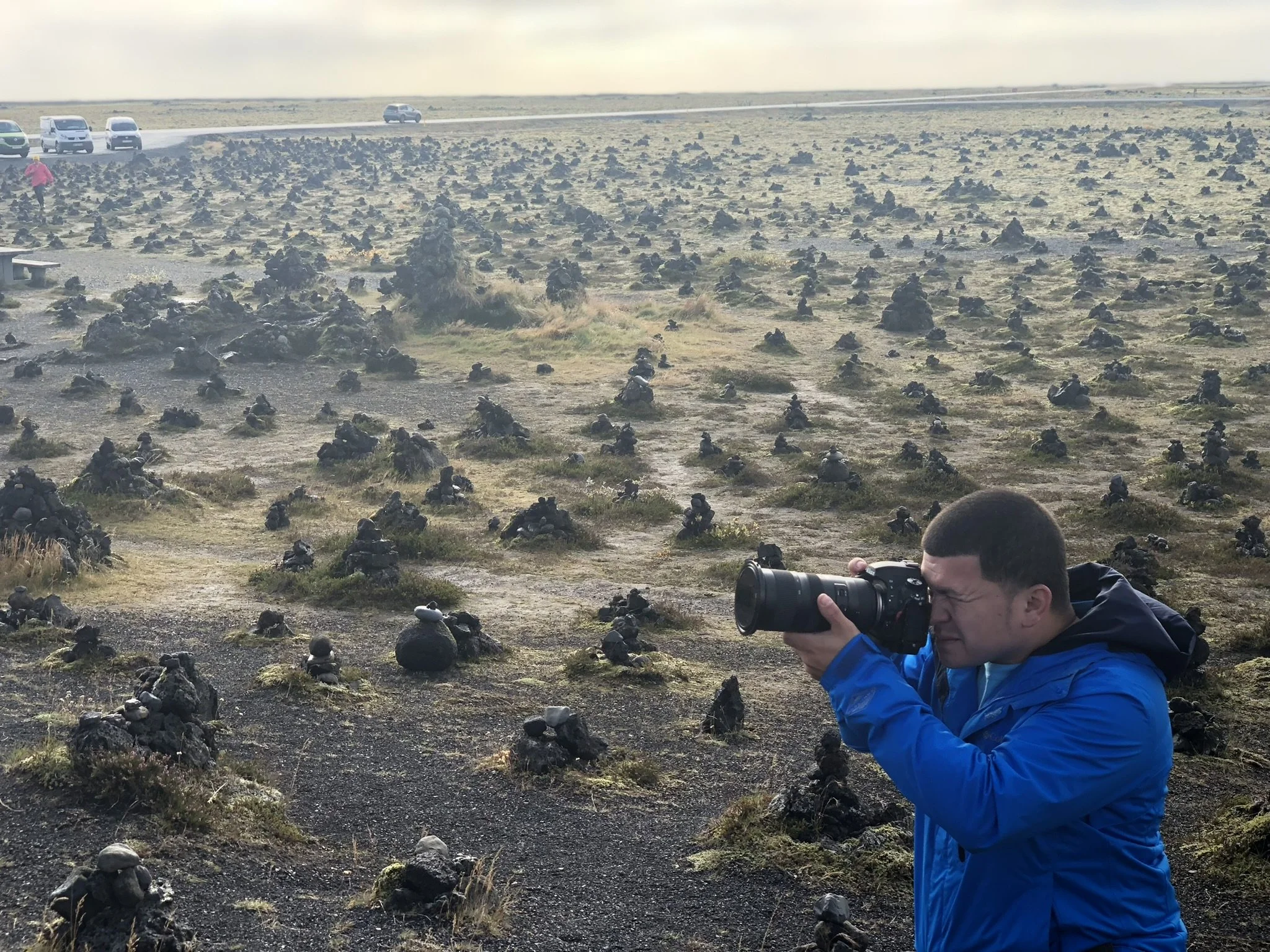 A young man in a blue jacket takes a photograph with a camera in a landscape filled with numerous black volcanic rocks and moss, with a road and several cars in the background.