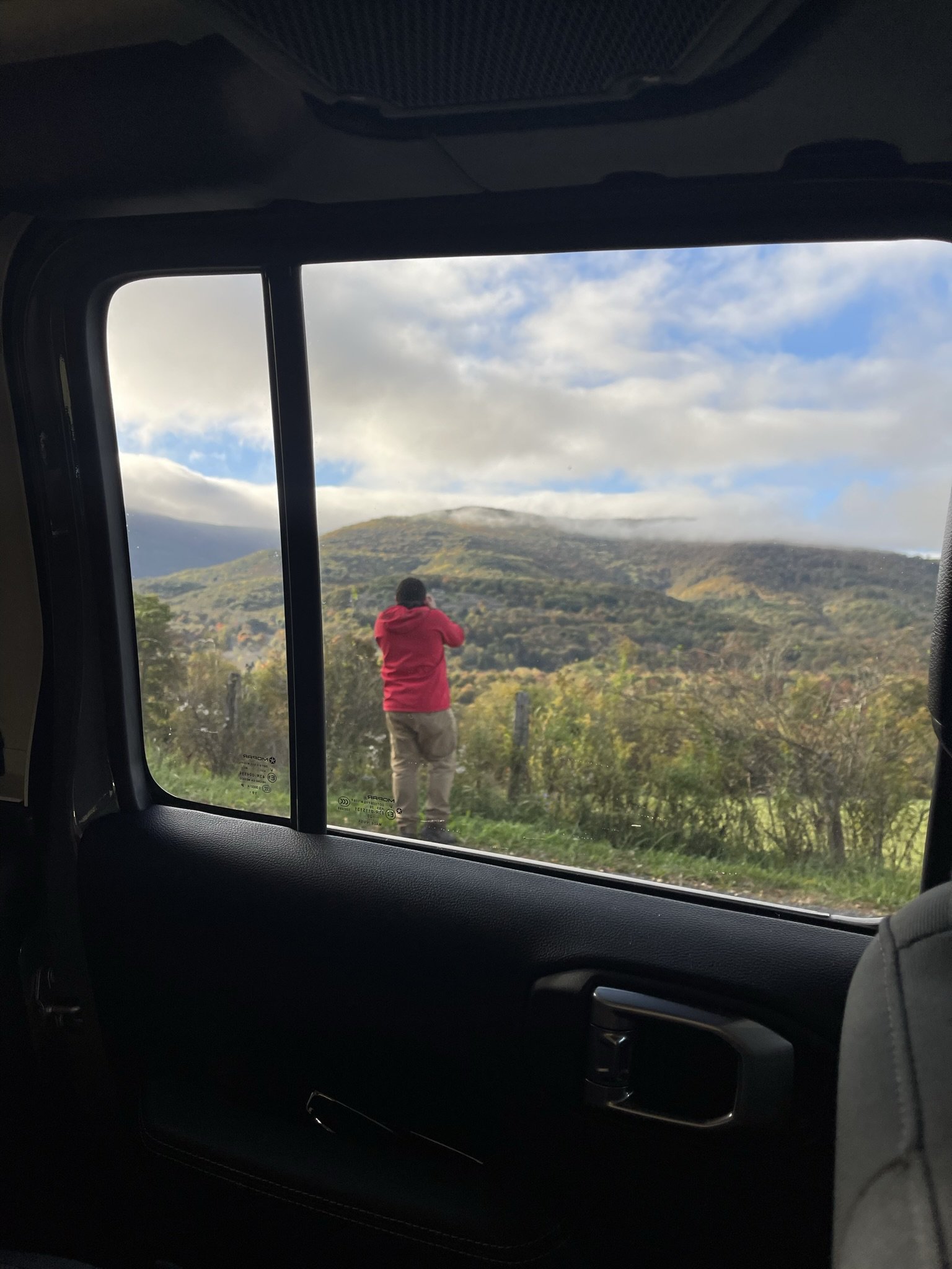 Person in a red jacket standing outdoors taking a photo of a mountain landscape with clouds and trees, viewed through a vehicle window.