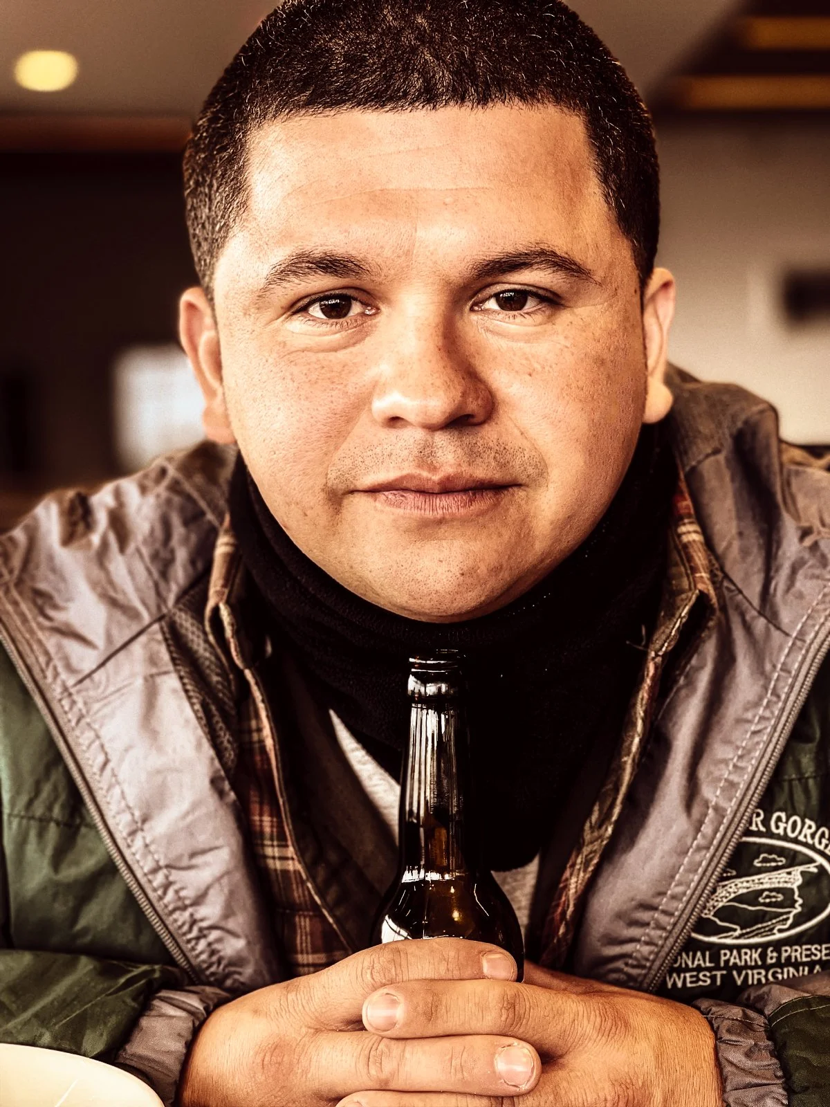 A man holds a brown glass bottle of beer, sitting indoors. He has short dark hair, wearing a layered jacket and a black scarf, with a slight smile on his face.