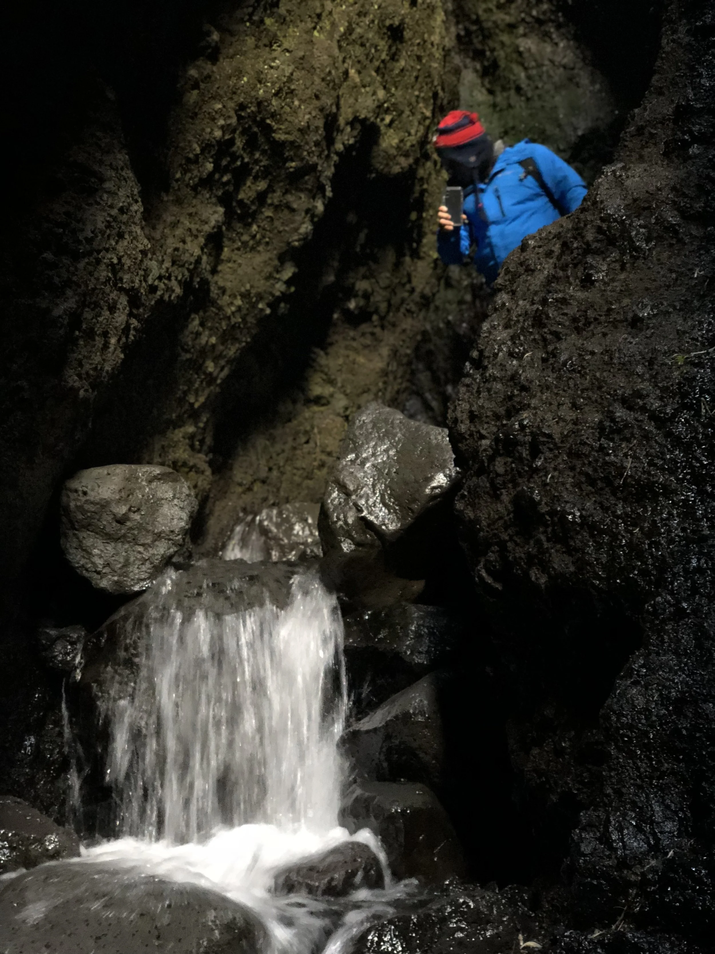 Person wearing a blue jacket and a red and black beanie, taking a photo with a smartphone inside a dark rocky cave with flowing water.