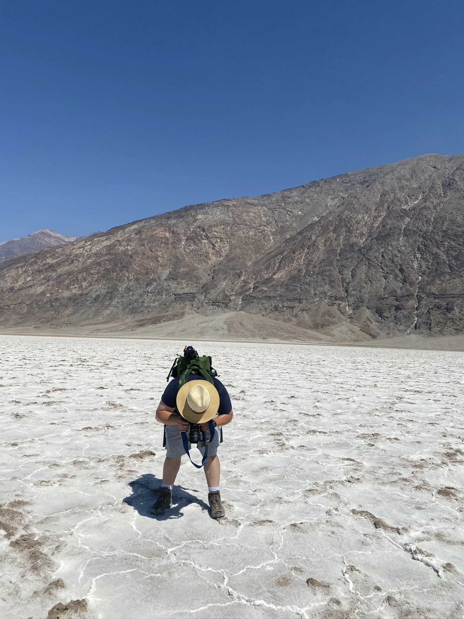Person in a hat and hiking gear holding a camera, standing on a white salt flat with mountains in the background and clear blue sky above.