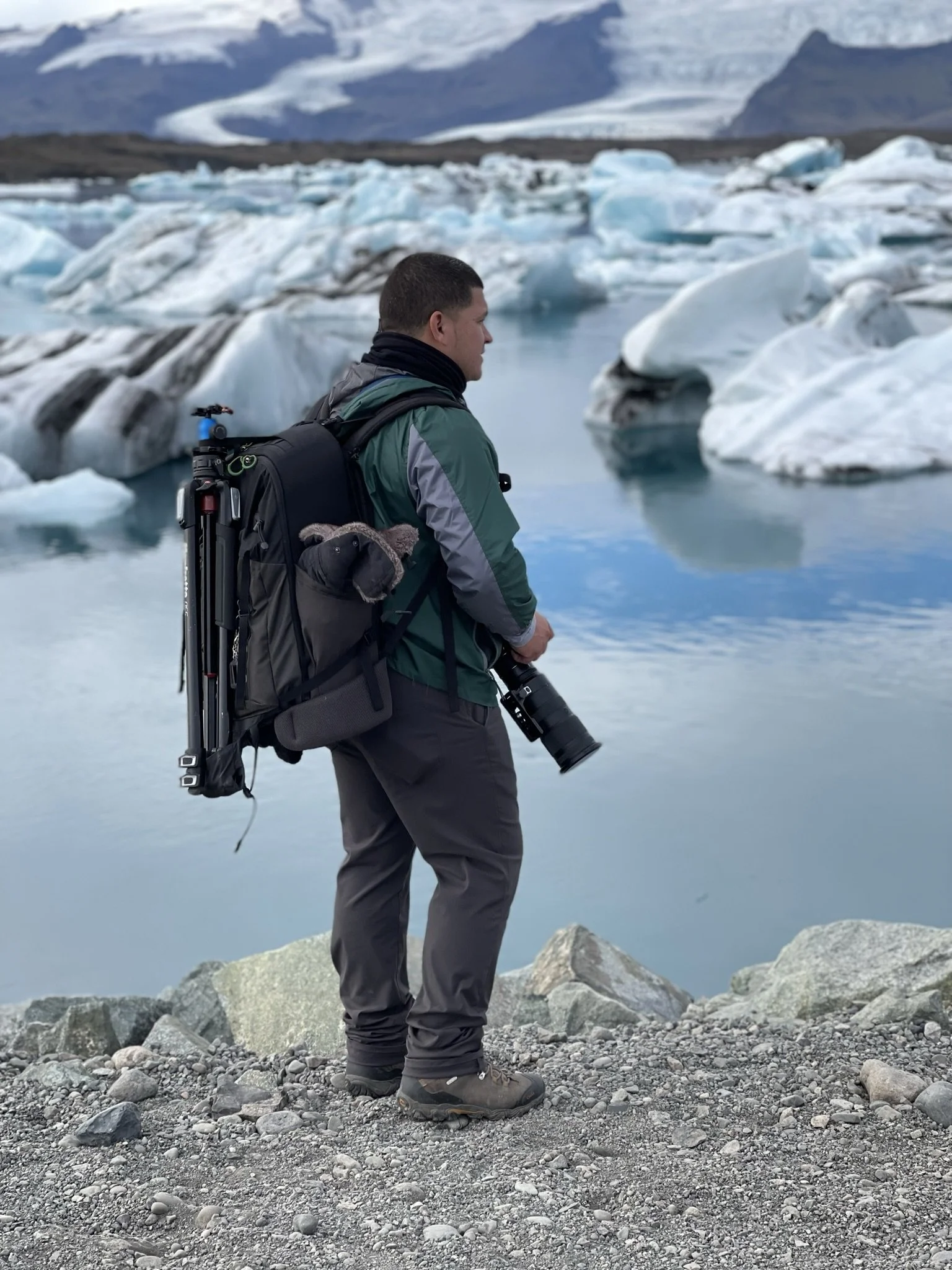 A man standing on rocky terrain near a glacial lake, holding a camera, with icebergs and mountains in the background.