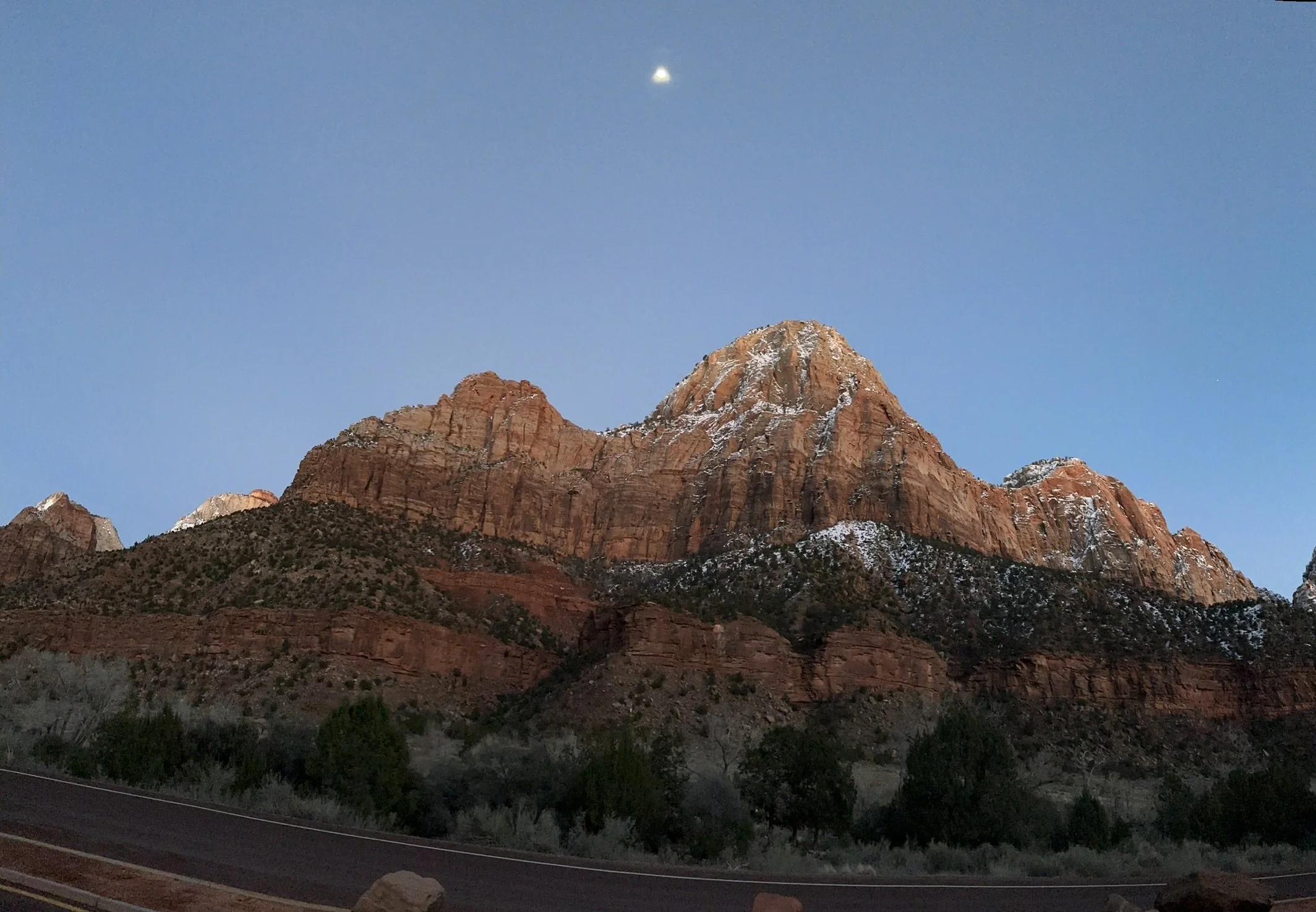 A mountain range with red rock formations and patches of snow, under a clear blue sky with visible moon.