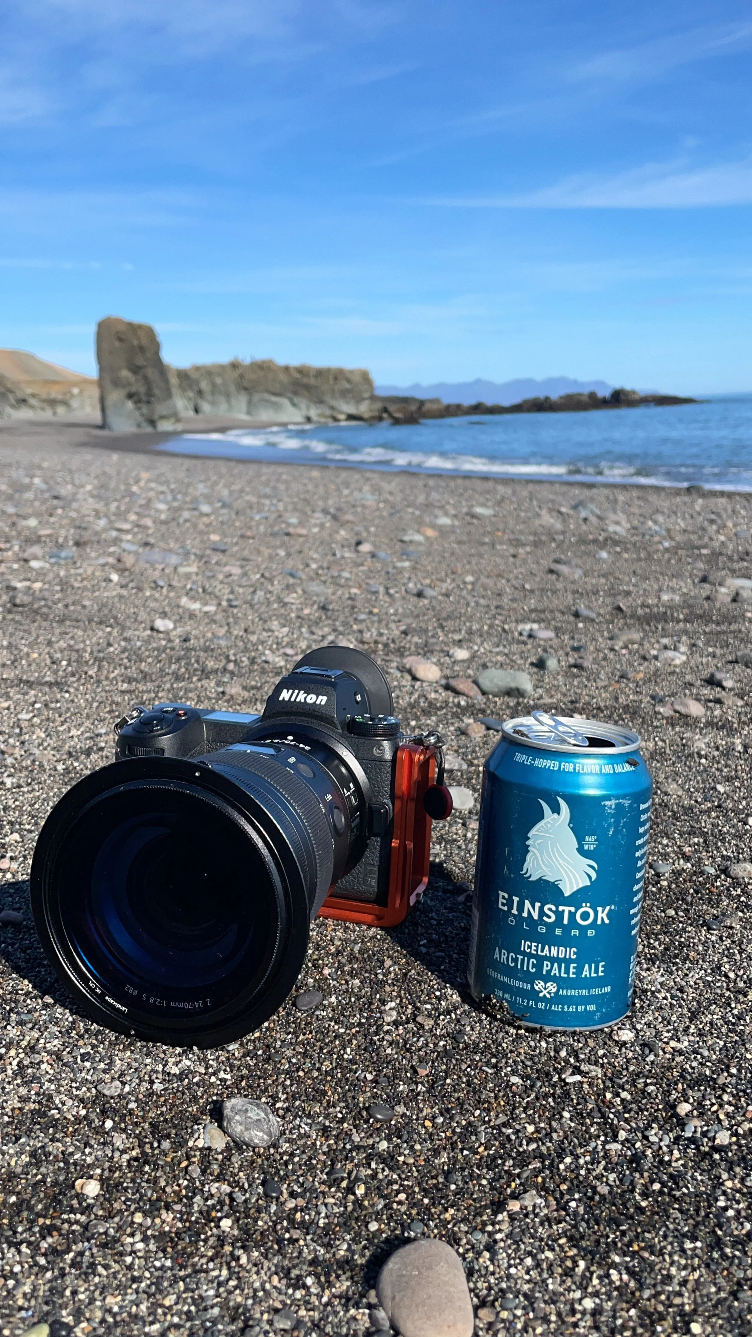 Camera and a can of Einstok Arctic Pale Ale on a pebbled beach with cliffs and the ocean in the background.